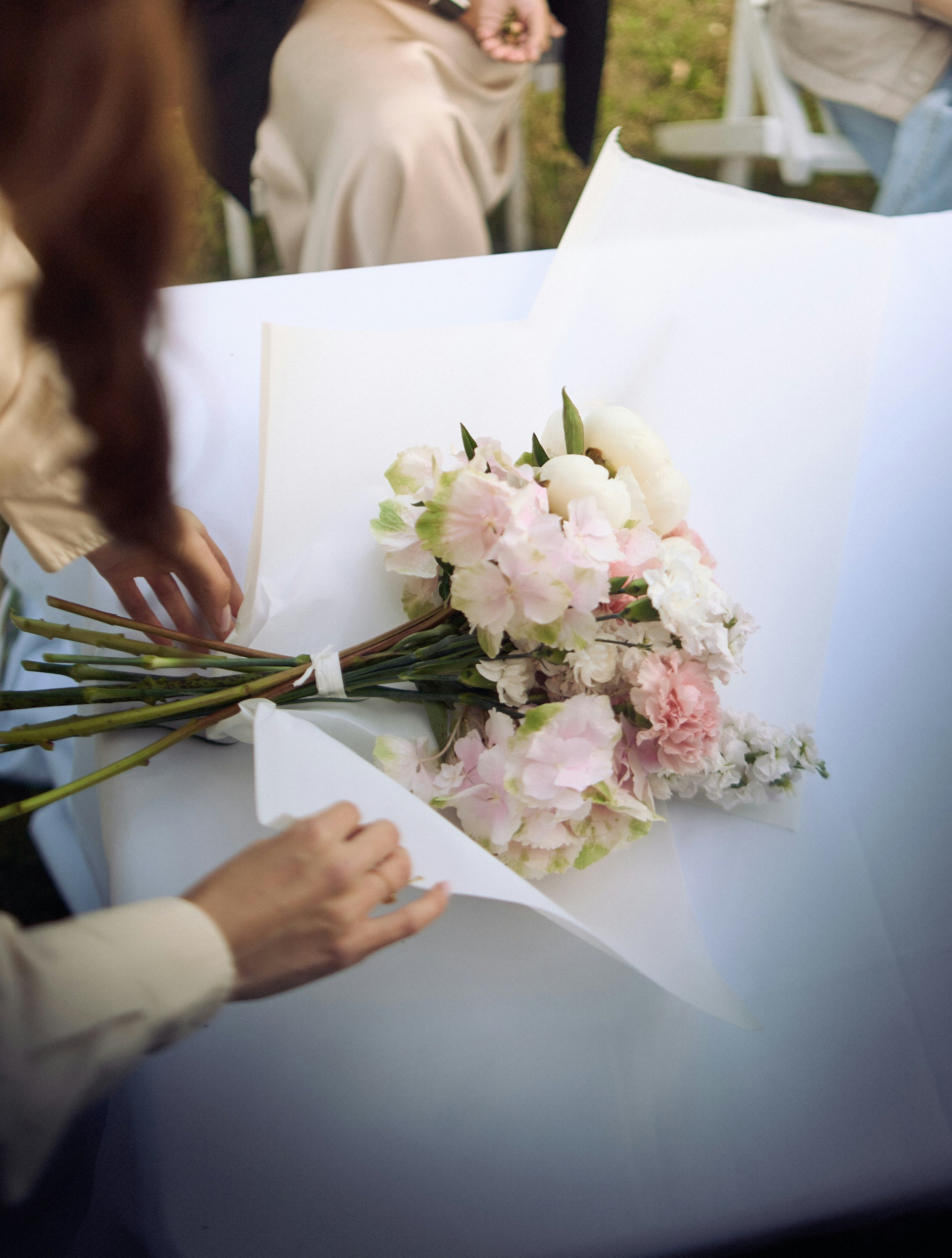 People wrapping pink and white flower bouquet in white paper outdoors.