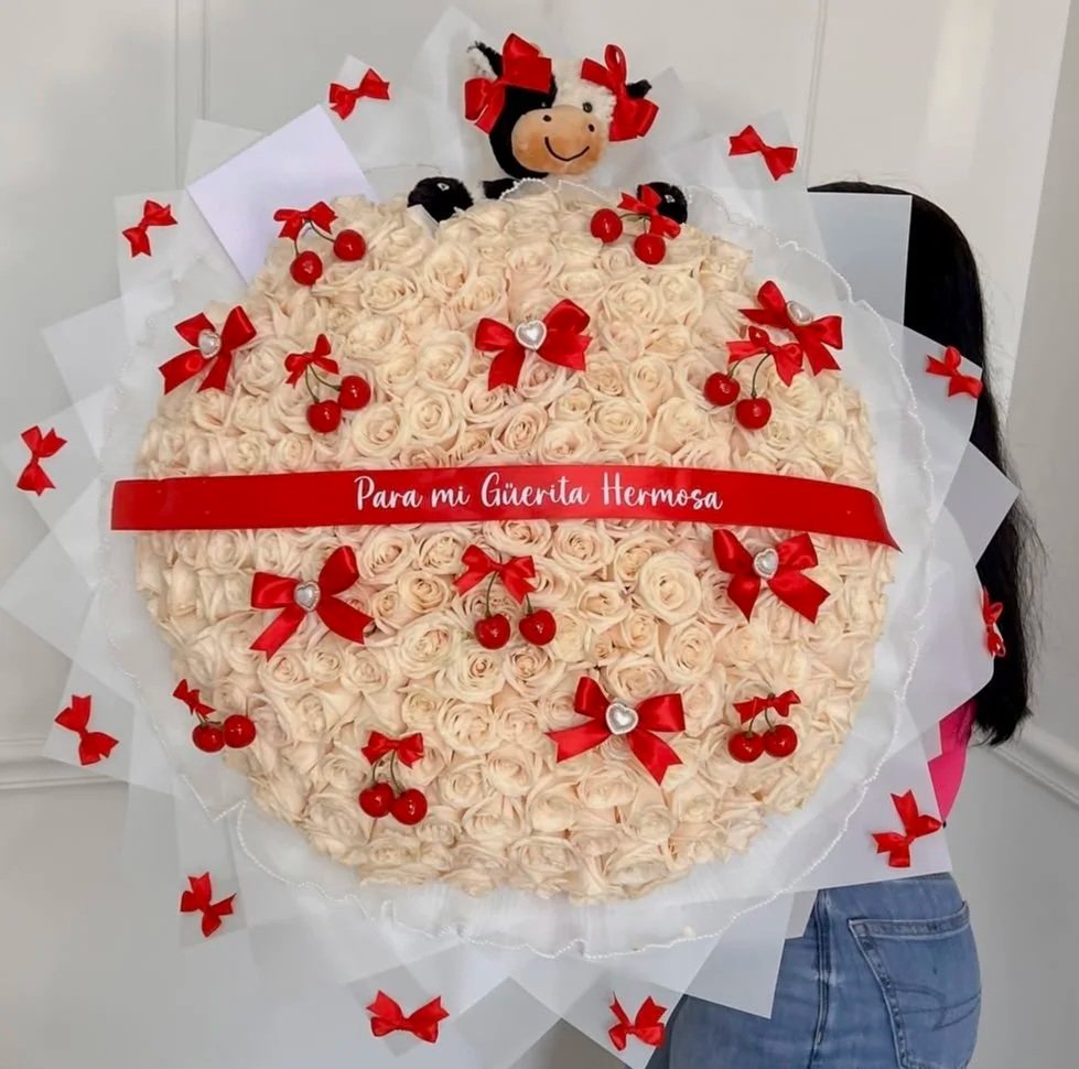 Person holding a large round flower bouquet decorated with small pink roses, red ribbons, cherries, and a plush doll at the top. The bouquet has a red ribbon with a message in Spanish that says "Para mi Gáuerrita Hermosa".