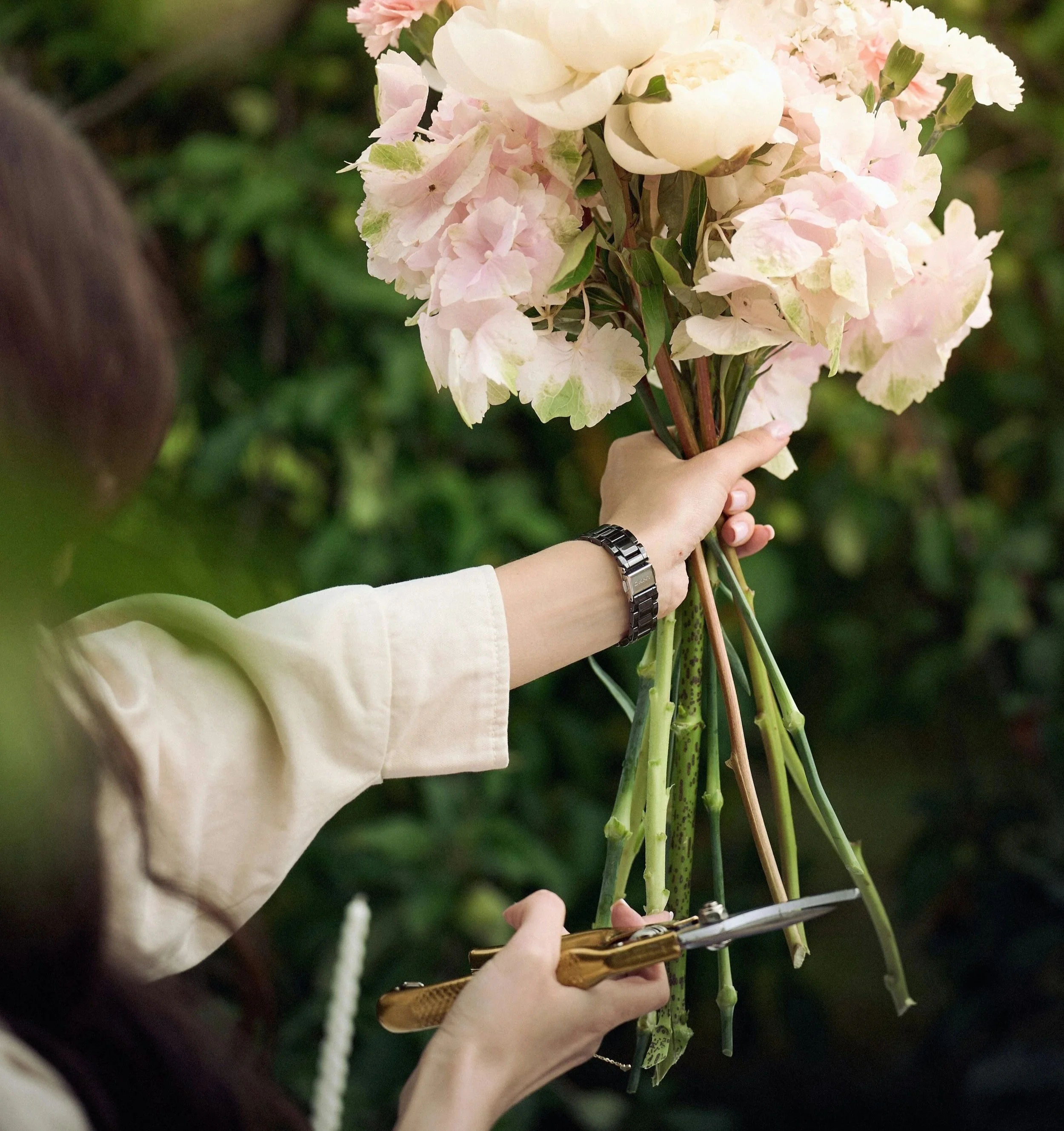 A person holding a bouquet of light pink and white hydrangea flowers with green leaves, against a blurred green background.