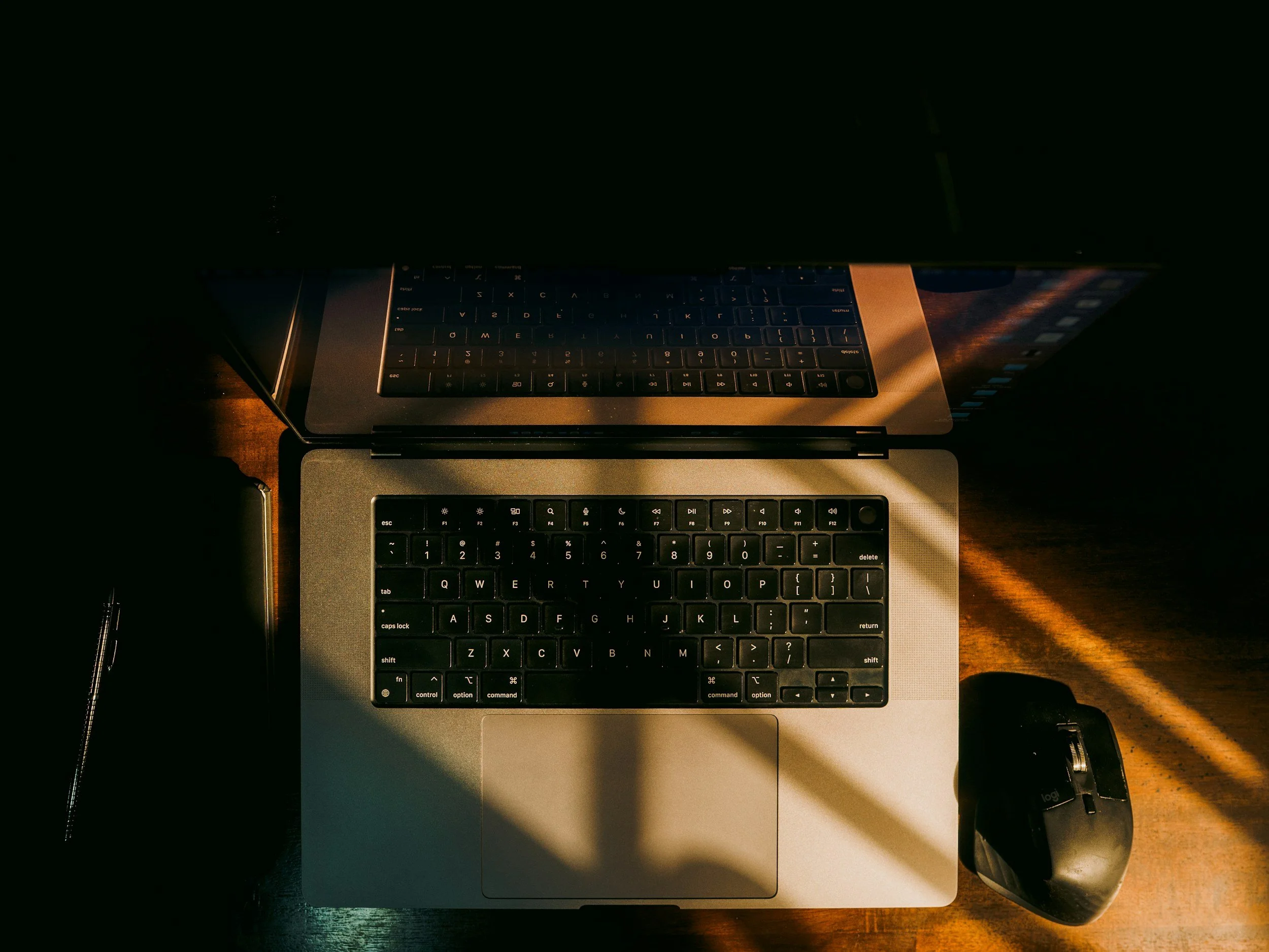 Overhead view of a MacBook laptop with a keyboard and trackpad, a wireless mouse, a closed notebook, and a pen on a wooden surface, illuminated by warm sunlight.