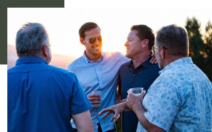 Four men outdoors at sunset, engaged in conversation and smiling, one wearing sunglasses, holding drinks, friends socializing.