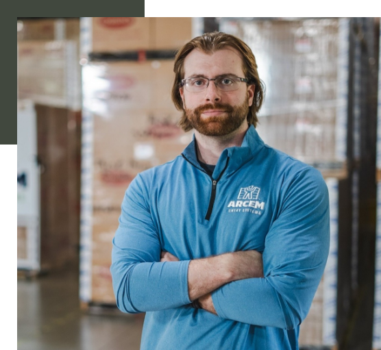 A man with glasses and a beard, wearing a blue Arcem jacket, standing with arms crossed in a warehouse with stacked boxes in the background.