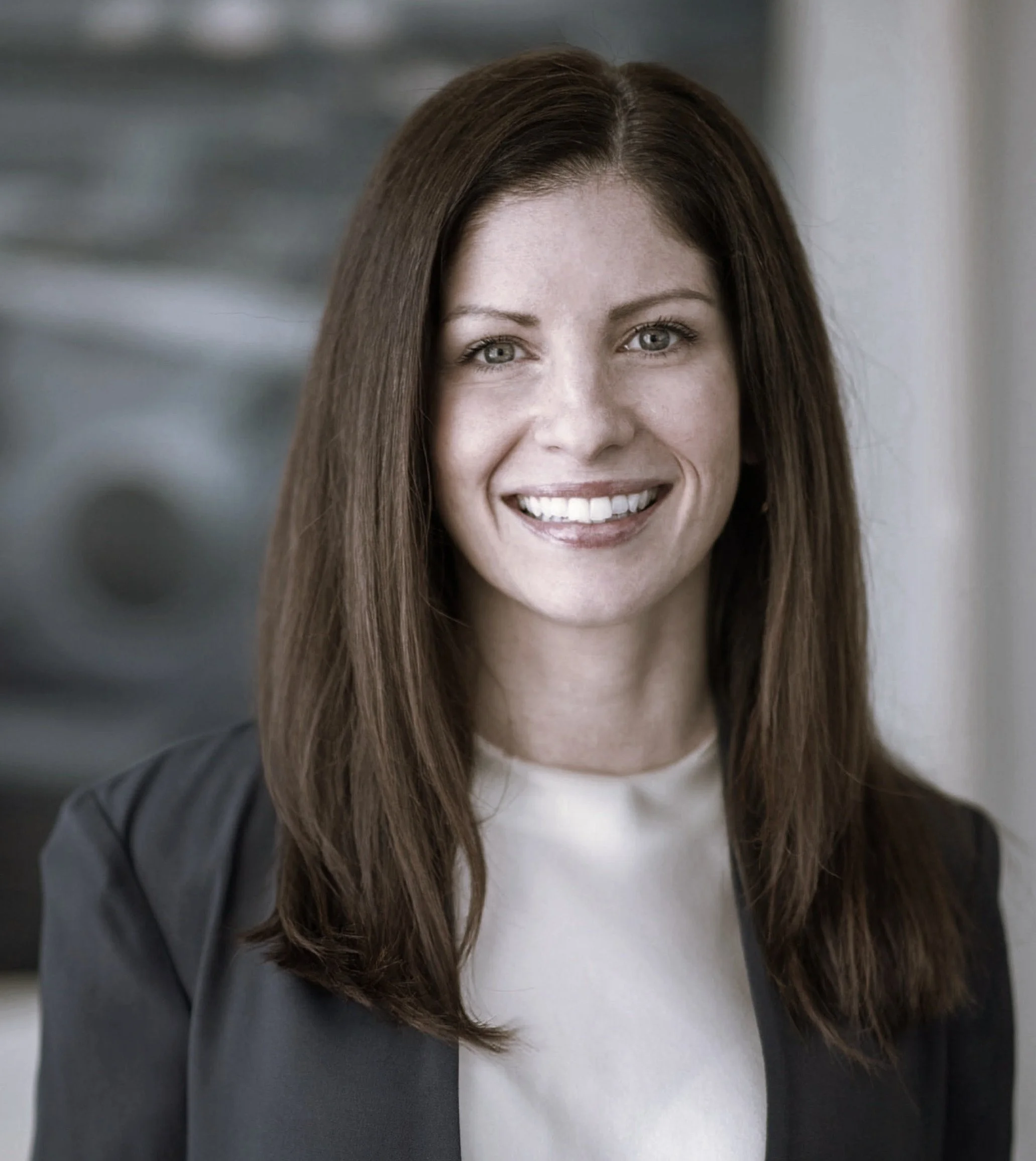 A woman with long brown hair, smiling, wearing a black blazer and white top, in an indoor setting.