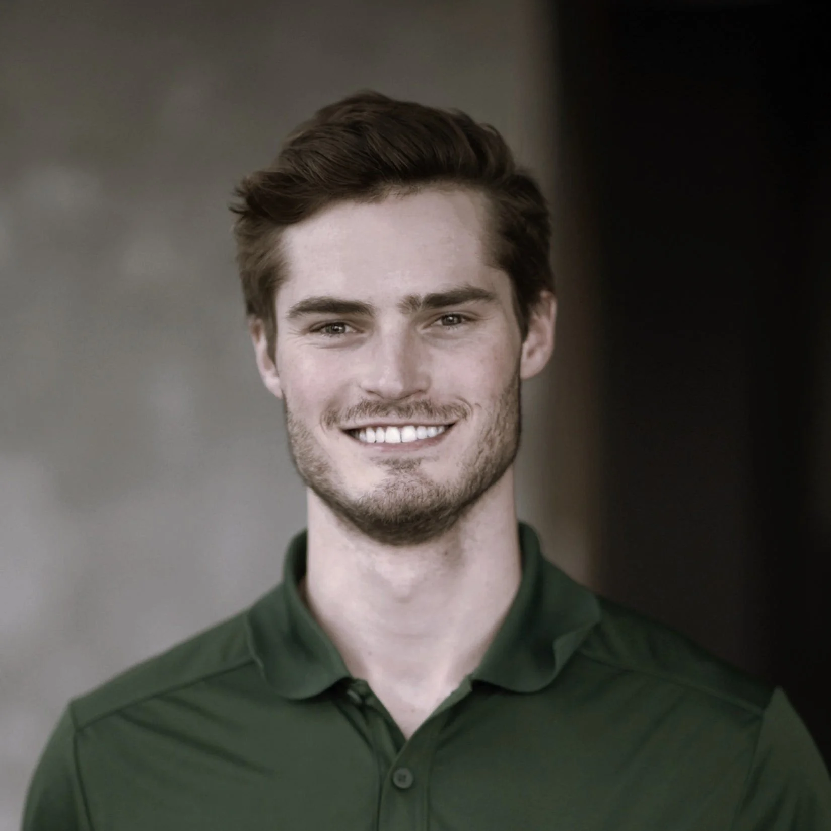 Young man with brown hair and a beard smiling, wearing a dark green polo shirt, outdoor setting