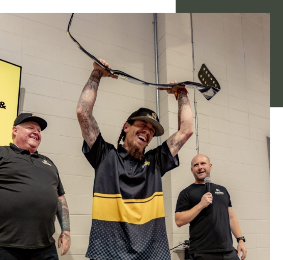 A man in a black and yellow polo shirt raises a hockey stick in the air, smiling, with two other men standing nearby; one is holding a microphone and wearing a black polo shirt, while the other is wearing a black baseball cap and shirt.