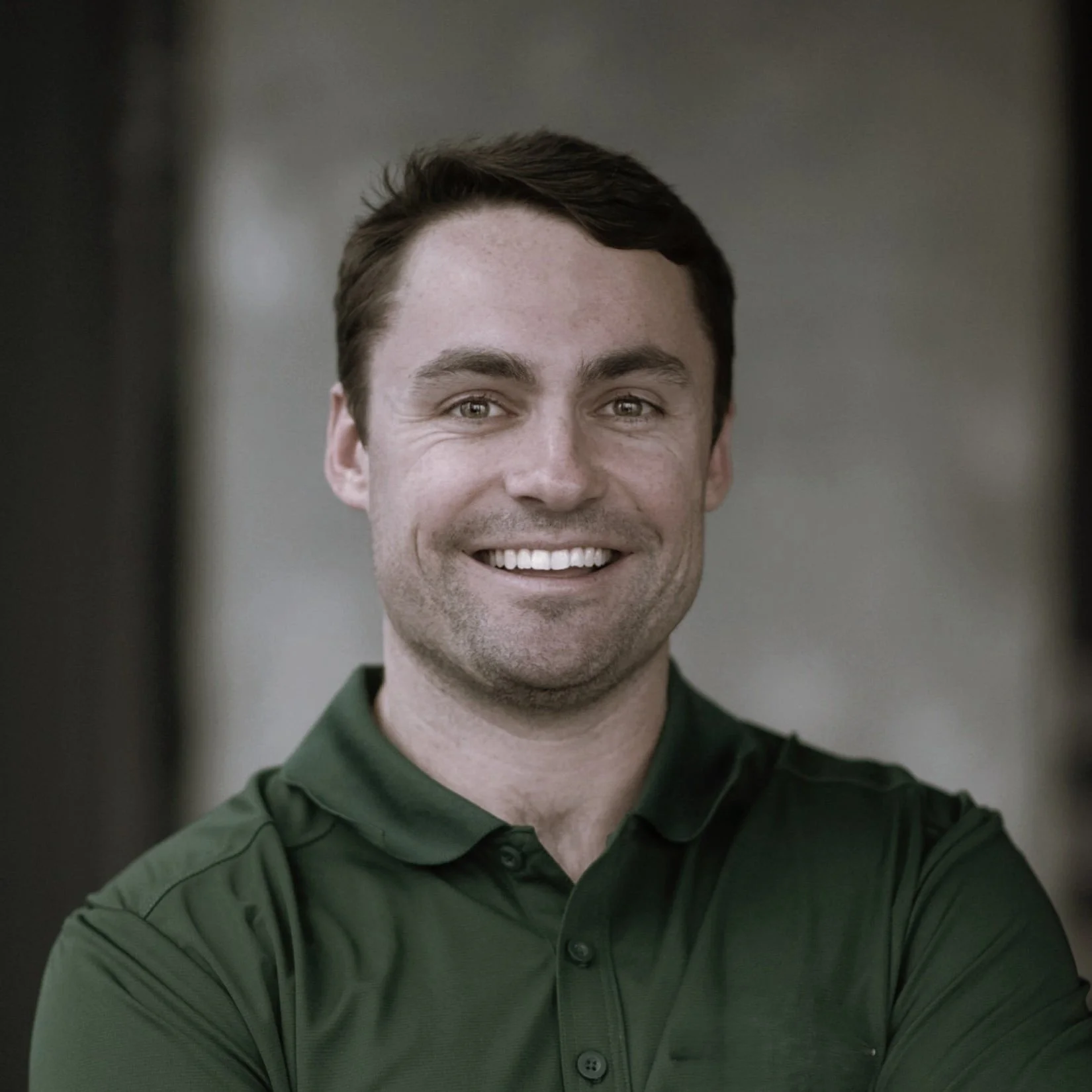 A smiling man wearing a dark green polo shirt, standing indoors with a blurred background.