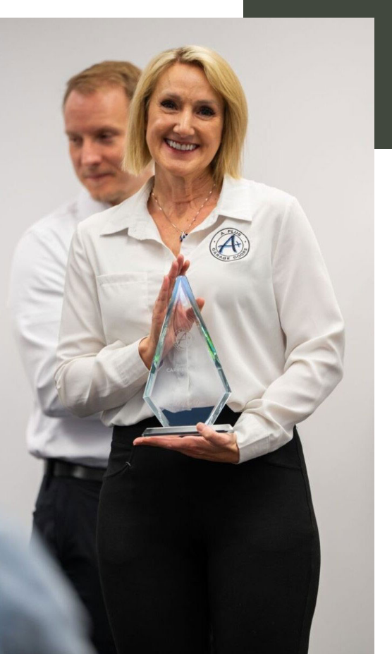 A woman with blonde hair, wearing a white button-up shirt and black pants, holding a glass award while smiling at the camera. A man in a white shirt is blurred in the background.