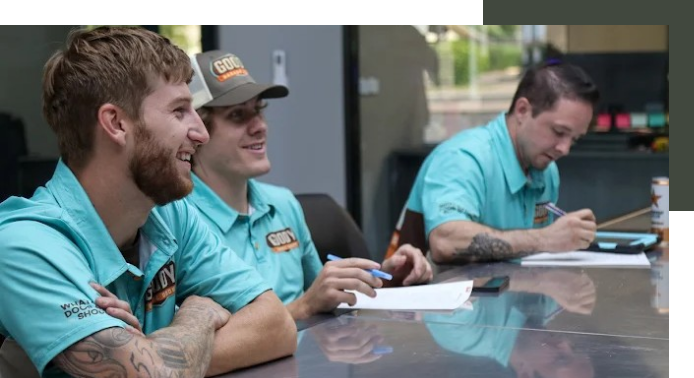 Three employees, wearing light blue shirts, sitting at a meeting table in a fast food restaurant, smiling and taking notes.