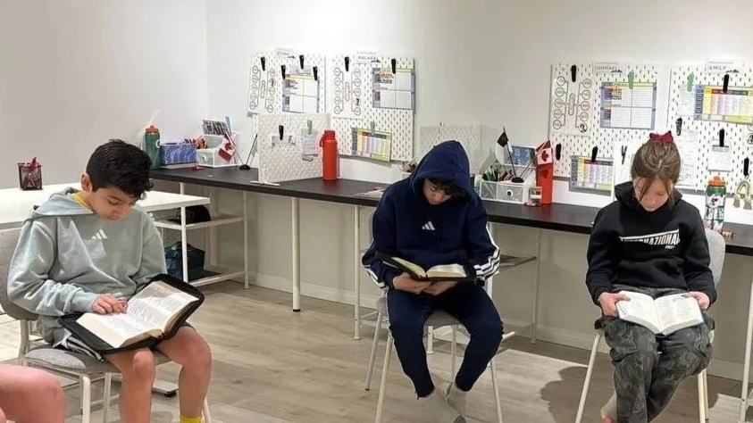 Three children sitting on chairs in a classroom, reading books. The classroom has white walls with chart posters and supplies on a long desk behind them.