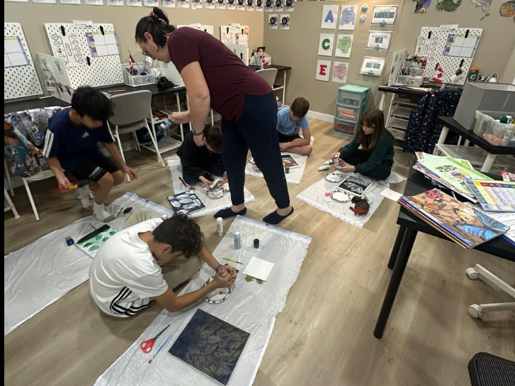 Children sitting on the floor in a classroom, engaging in art projects, with a teacher supervising