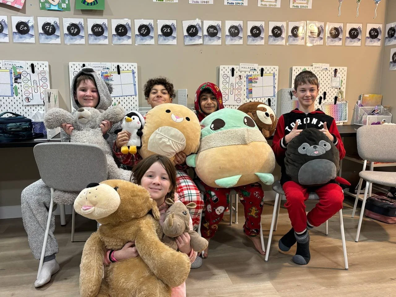 Five children sitting and kneeling, all holding large plush animal pillows in a classroom setting with schoolwork on the wall behind them.