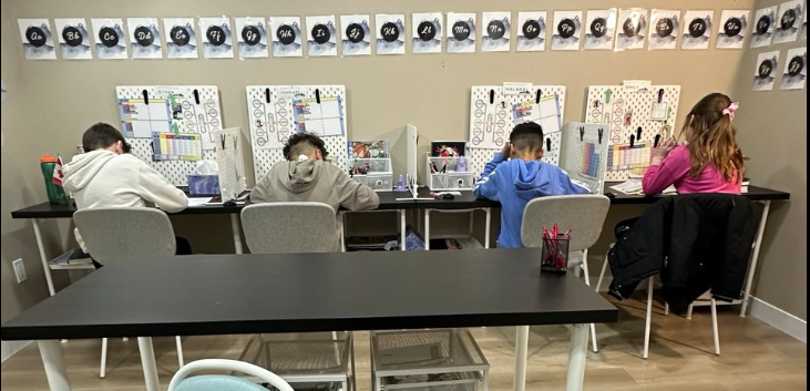 Four children sitting at desks in a classroom, working on assignments with educational charts and materials on the wall behind them.