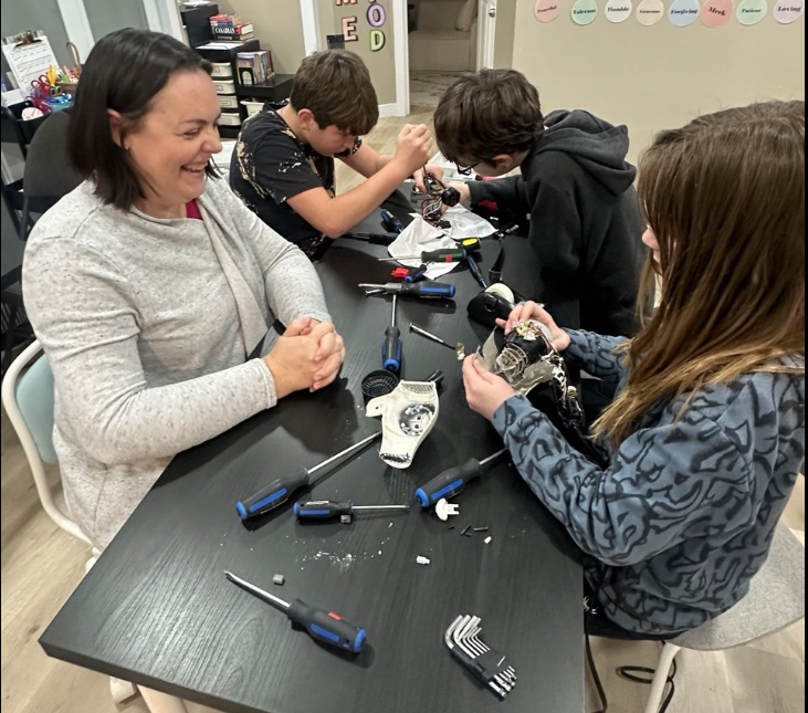 A woman and three children working together at a table on a mechanical project, with various screwdrivers and small parts on the table.