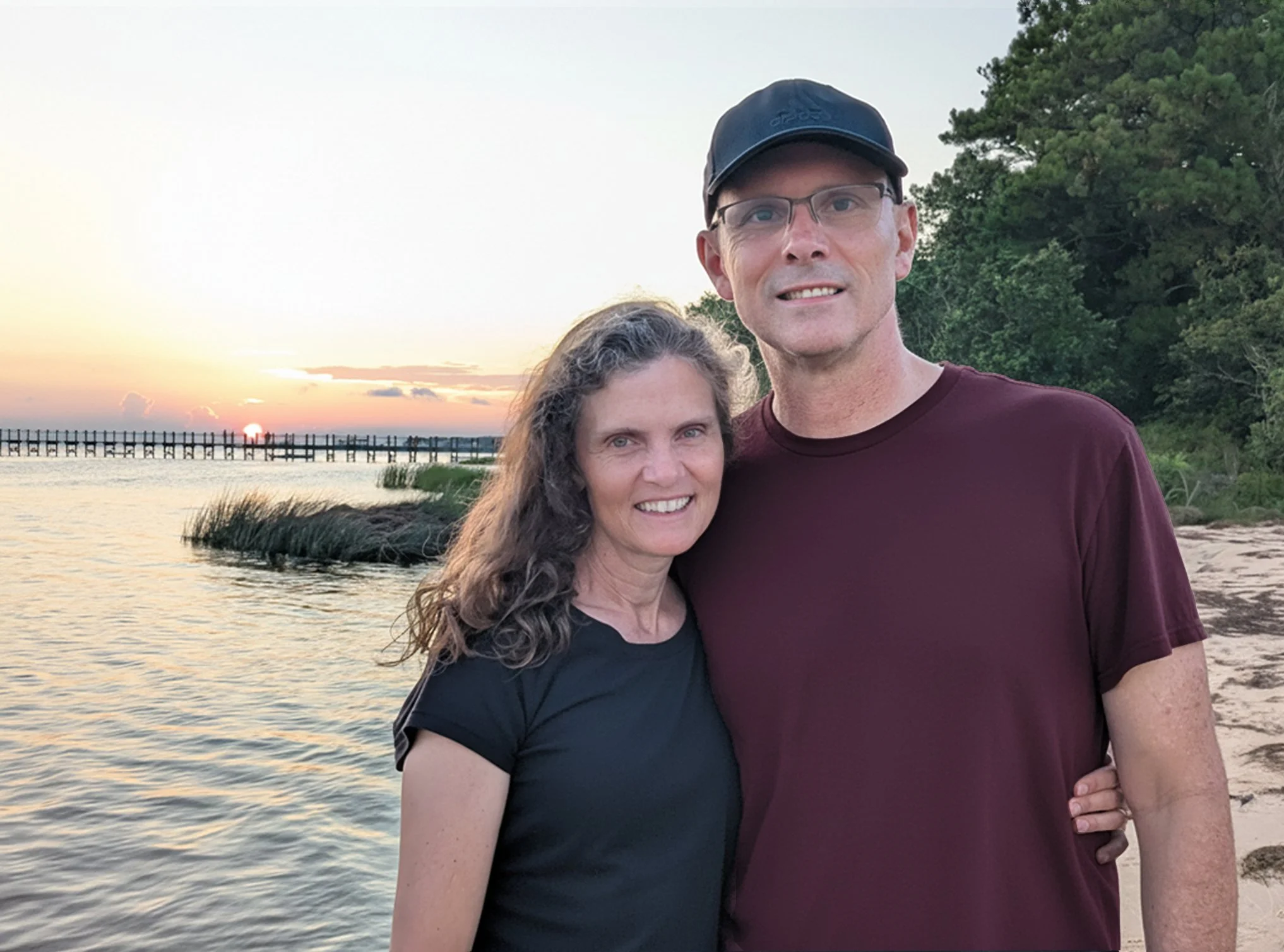 A woman and a man stand together on a beach during sunset, with the woman smiling and the man wearing glasses and a black cap, both surrounded by trees and water.