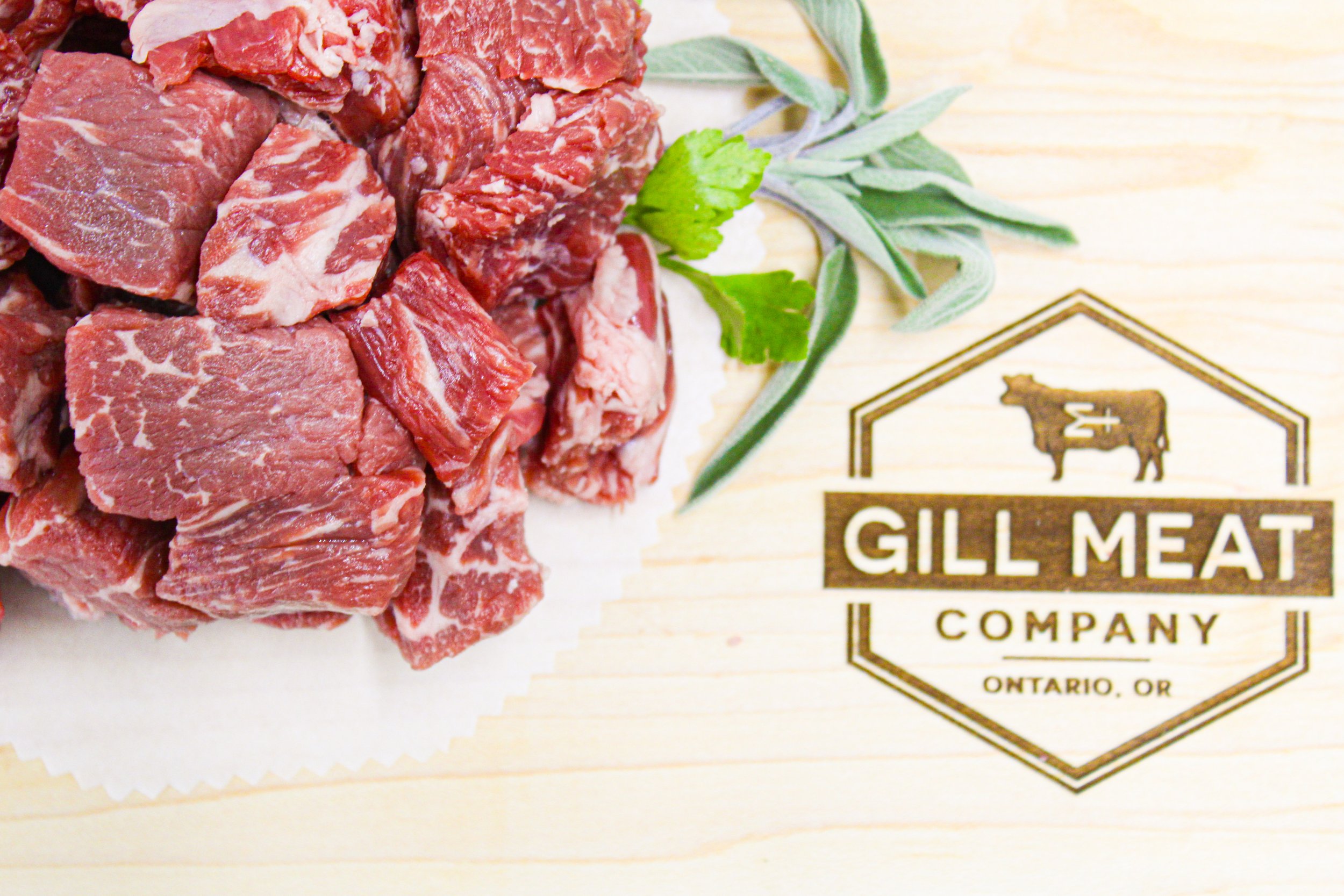 Fresh raw beef stew meat cubes from local pasture raised beef on a cutting board with green herbs and a GILL MEAT COMPANY logo.