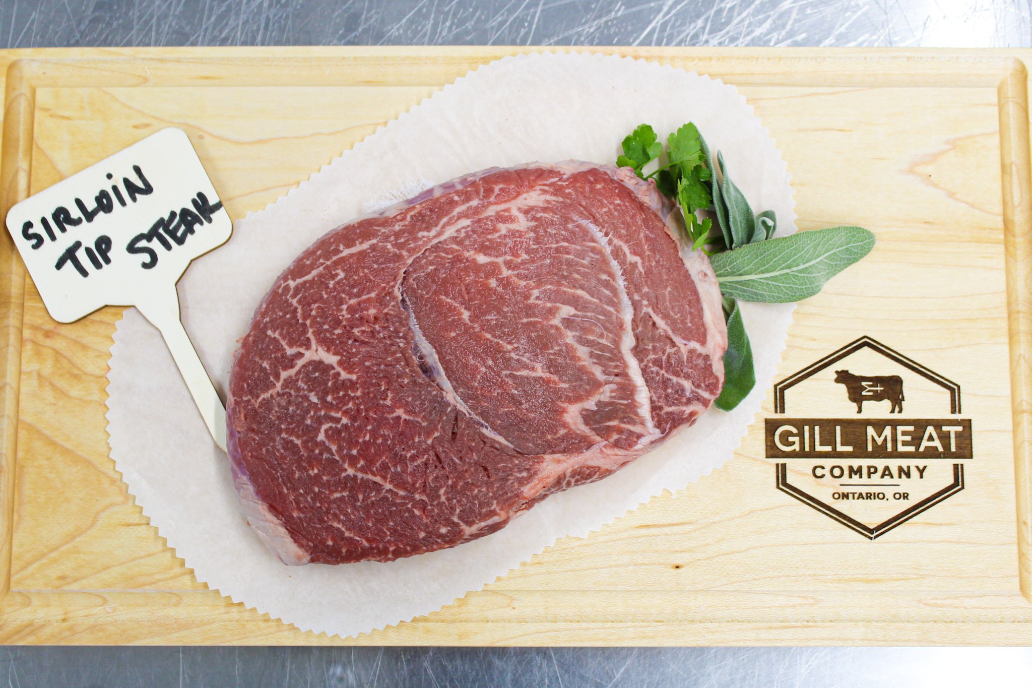 Raw sirloin tip steak on a wooden cutting board garnished with herbs, with a label indicating recent butchering, and the logo of Gill Meat Company from Ontario, Oregon.