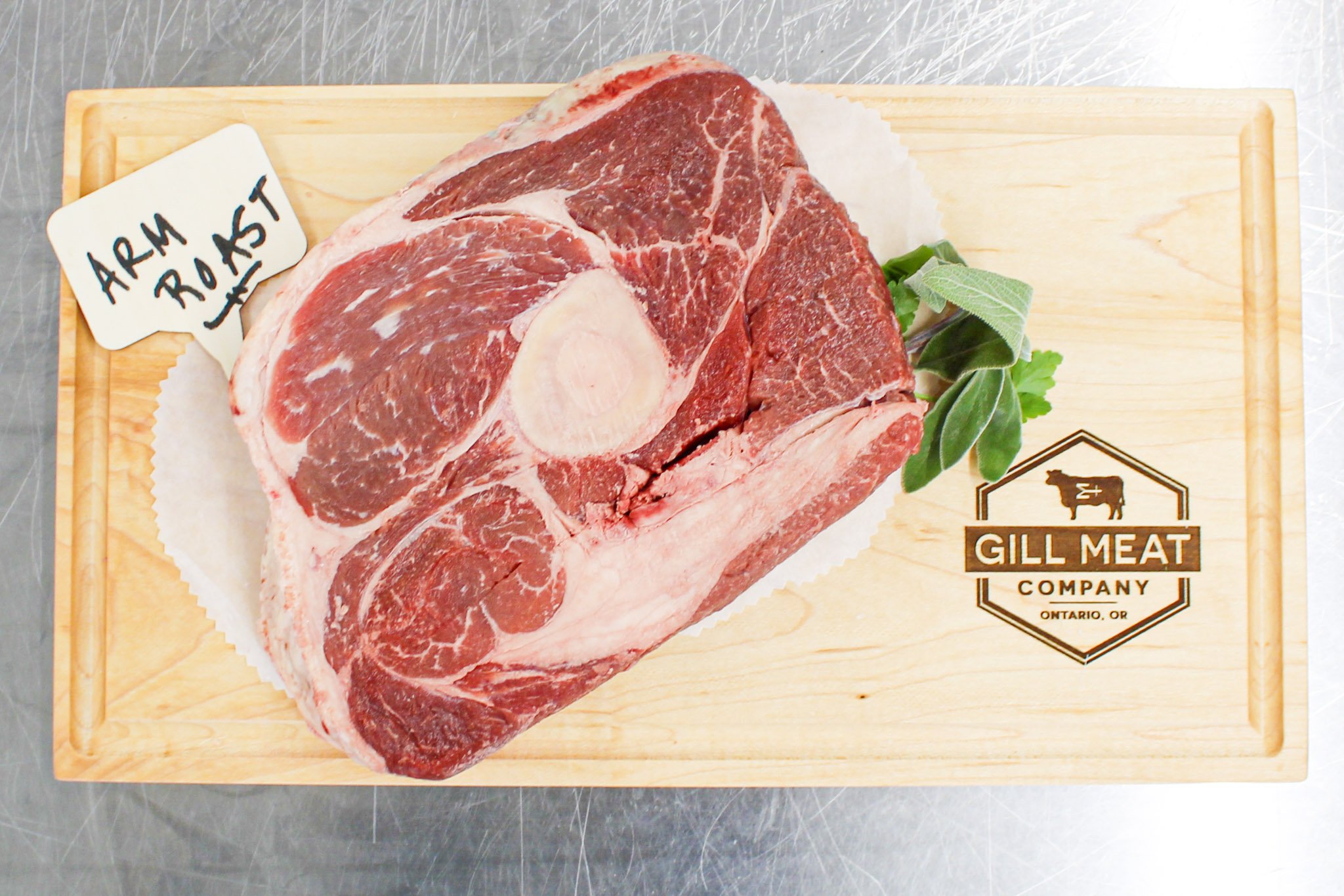 A large piece of red marbeled raw arm roast beef from pasture raised local beef on a wooden cutting board with a small sign that says 'ARM ROAST' and some green herbs. The cutting board has a logo for Gill Meat Company, Ontario, OR.