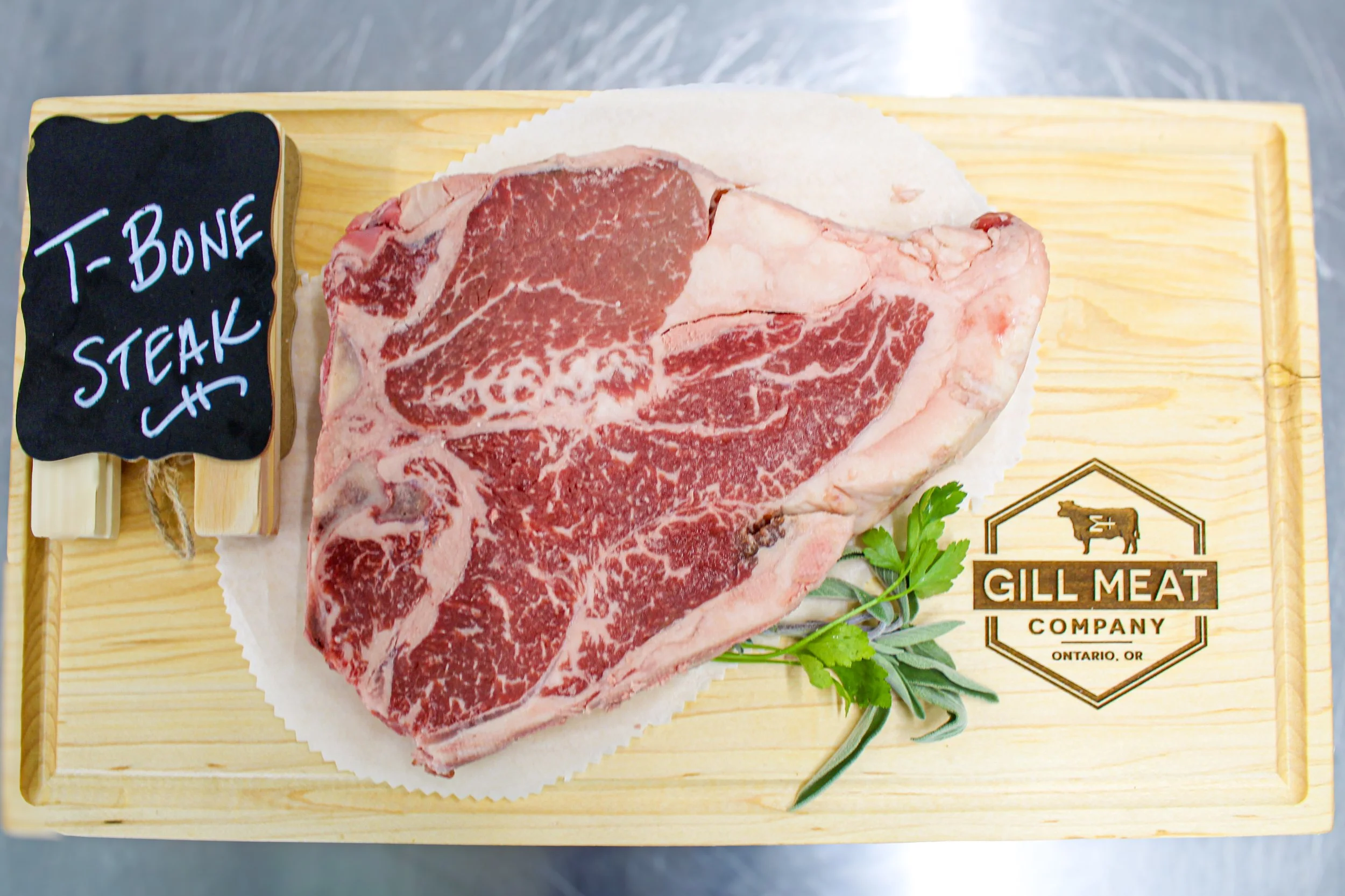 Raw well marbled grass fed corn finished T-bone steak on a wooden butcher's board with a small chalkboard sign reading 'T-Bone Steak' and a garnish of parsley and sage, with the Gill Meat Company logo from Ontario, OR.