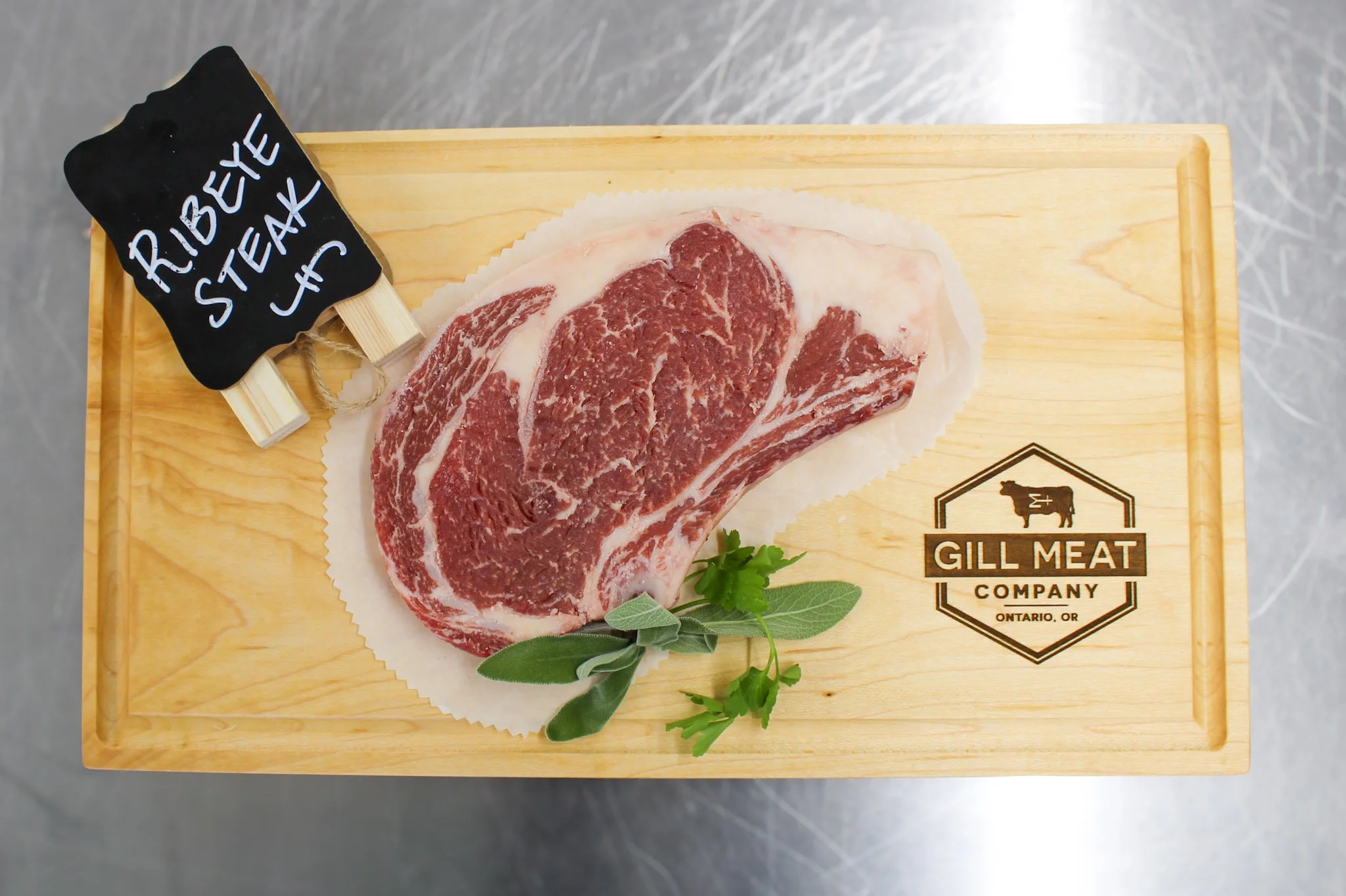 A raw ribeye steak on a wooden cutting board with a small black chalkboard sign labeled 'RIB EYE STEAK' and some green herbs.