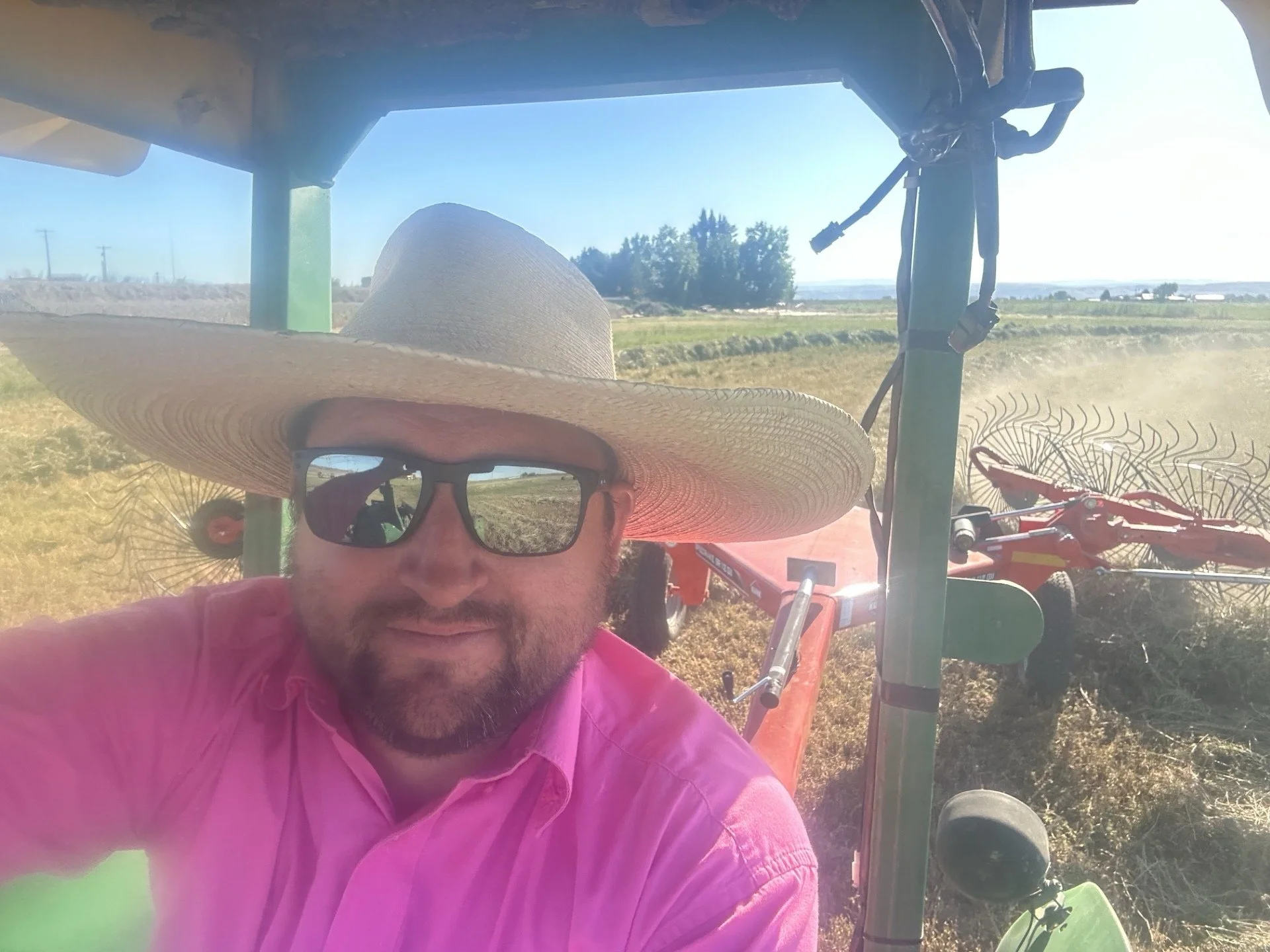 6th generation rancher from 140 year old ranching family man with a beard wearing sunglasses, a large straw hat, and a pink shirt, taking a selfie on a tractor haying alfalfa bales to feed to their pasture raised cattle. 