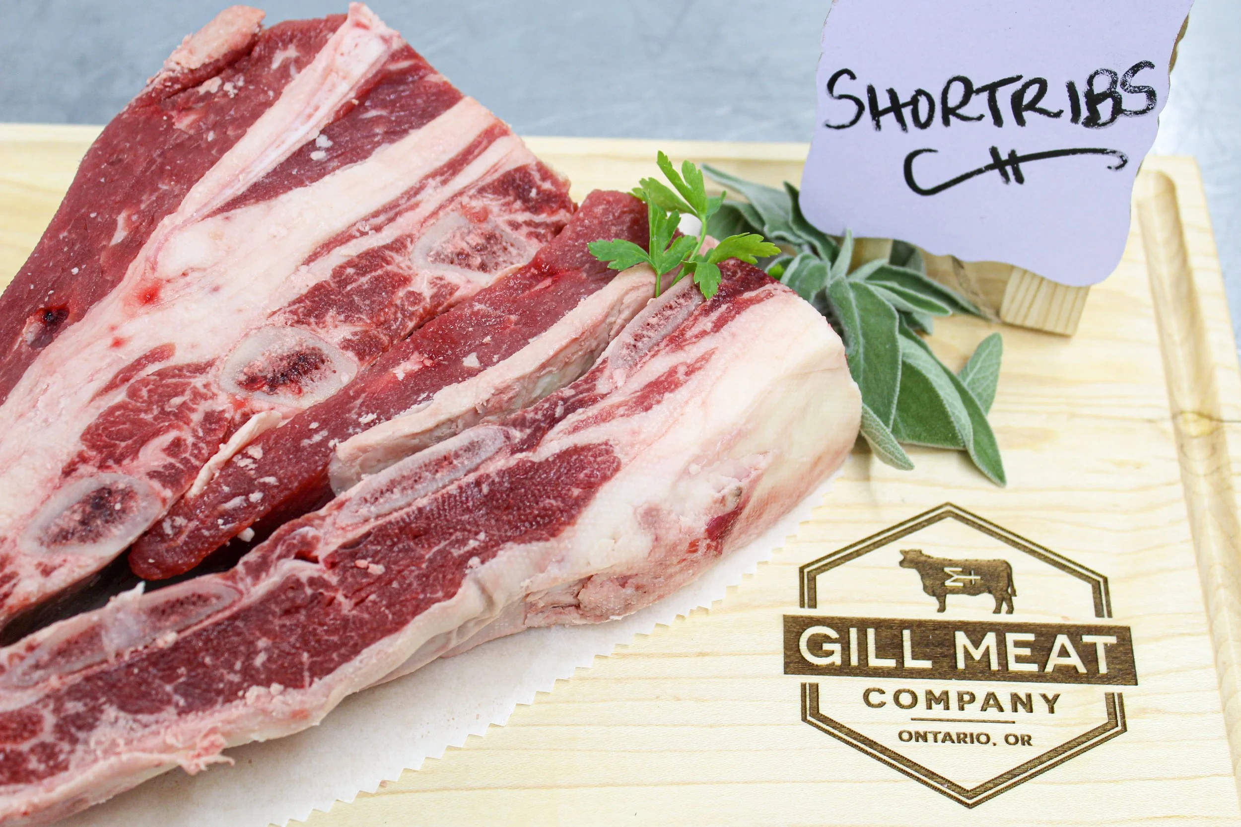 Fresh beef short ribs from red juicy corn finished local beef on a wooden board with a paper label saying "Short Ribs" and the Gill Meat Company logo.