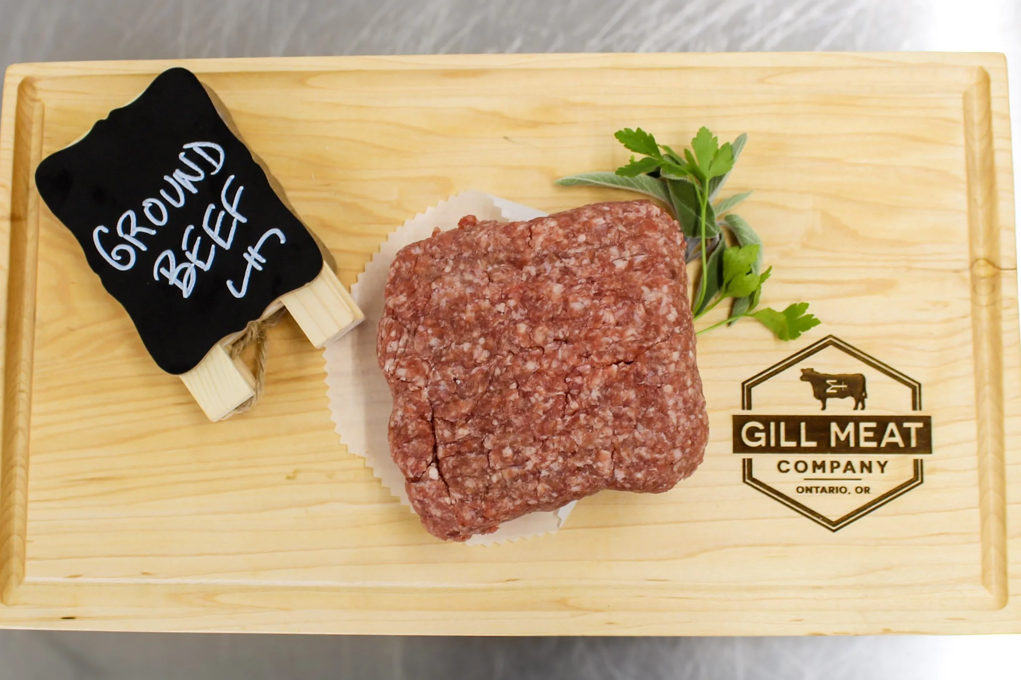 Raw ground beef patty on a wooden cutting board with a sprig of herbs, a small sign saying "GROUND BEEF," and a logo for Gill Meat Company on Ontario, Oregon.