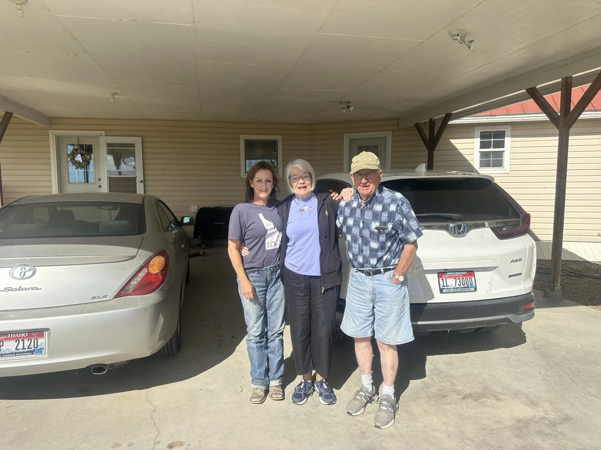 Three people from multi generational ranch and farm in boise area standing together in a driveway with two cars and a house behind them. The person on the left is a woman with brown hair, wearing a gray t-shirt and jeans. The person in the middle is 