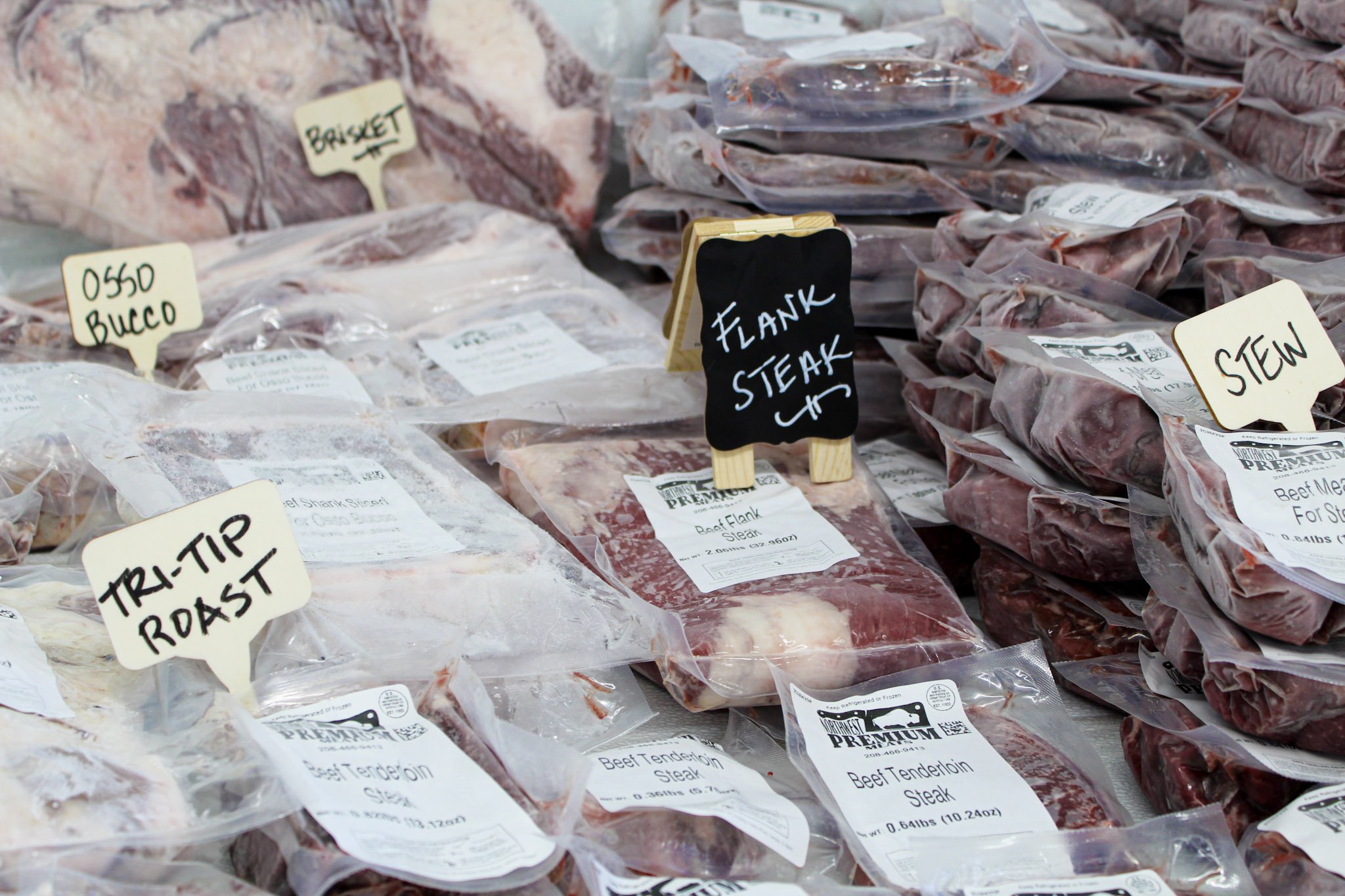 Various packages of raw beef cuts, including brisket, tri-tip roast, flank steak, and stew meat labeled and displayed on a table in a butcher shop with local fresh Oregon and idaho beef.