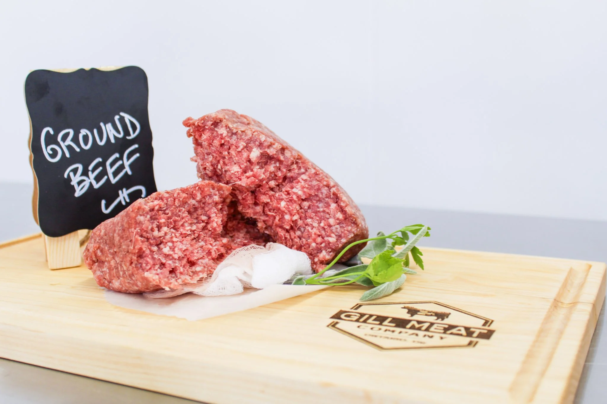 Raw ground beef on a wooden cutting board with a small black chalkboard sign labeled 'Ground Beef' and a sprig of green herbs.
