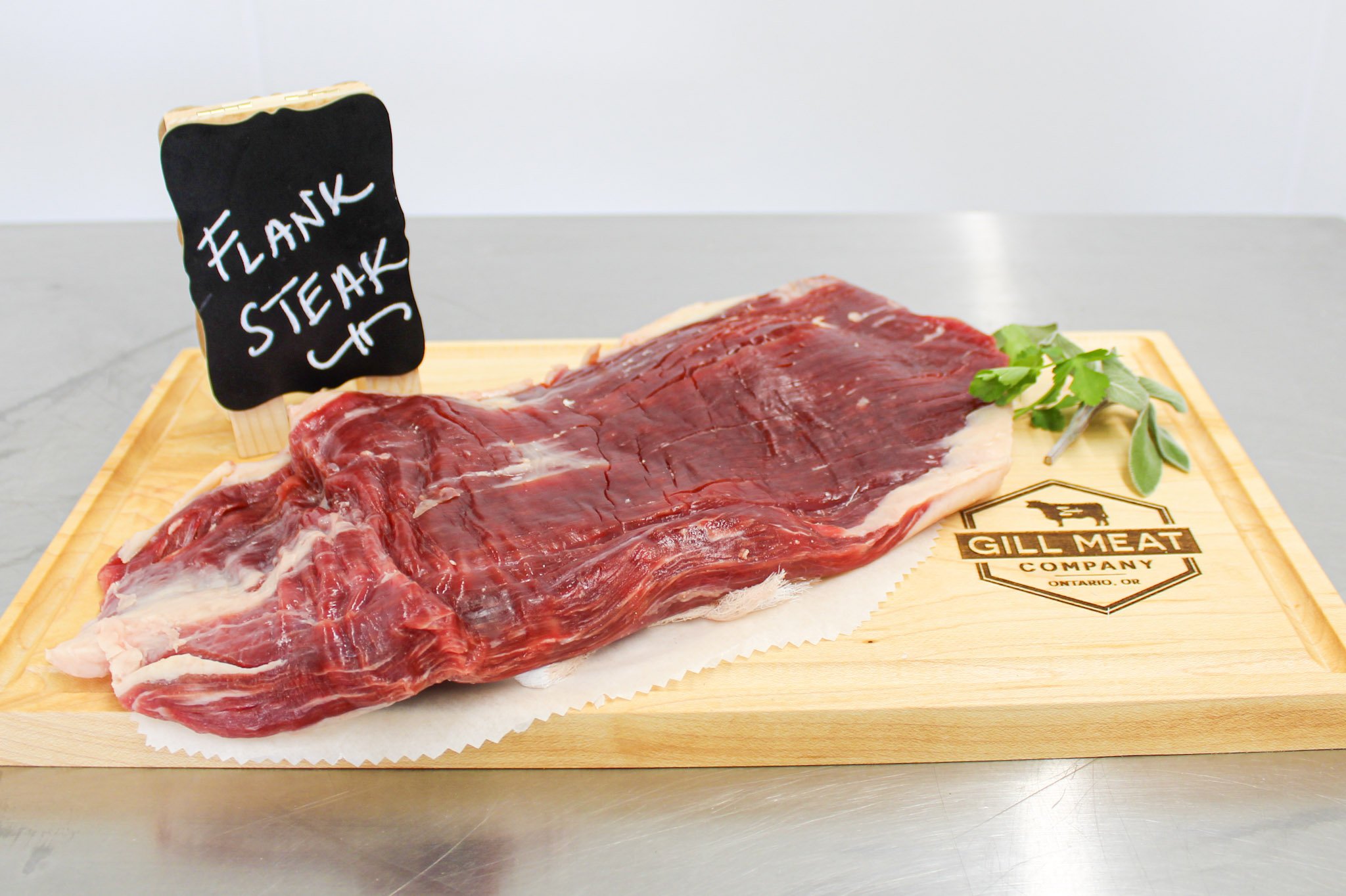 Raw flank steak on a wooden cutting board with a small chalkboard sign that says 'Flank Steak' and a sprig of herbs.