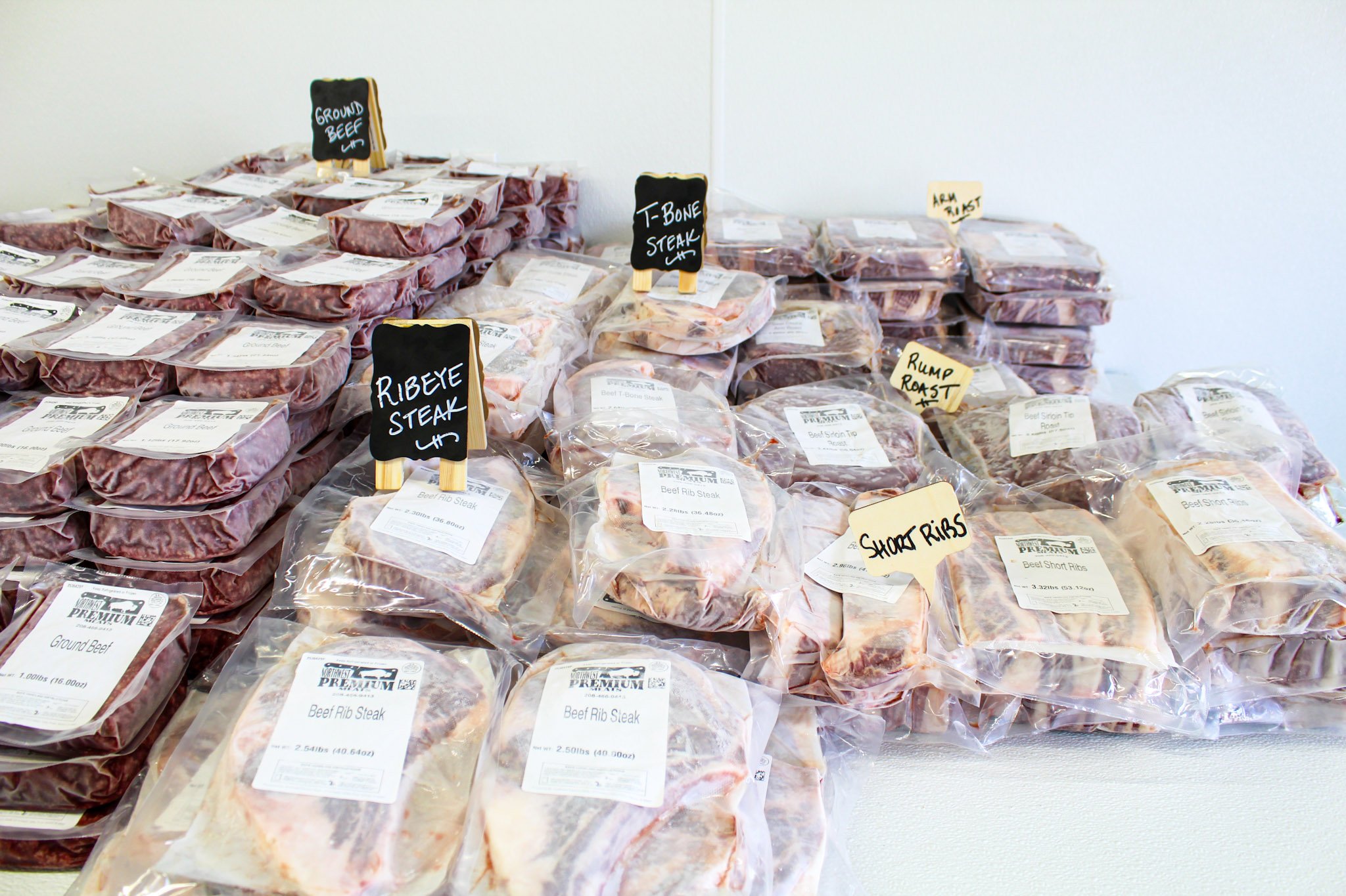 A display of various packaged beef cuts on a table, including ground beef, ribeye steak, T-bone steak, short ribs, and rump roast, with small handwritten labels indicating each cut.