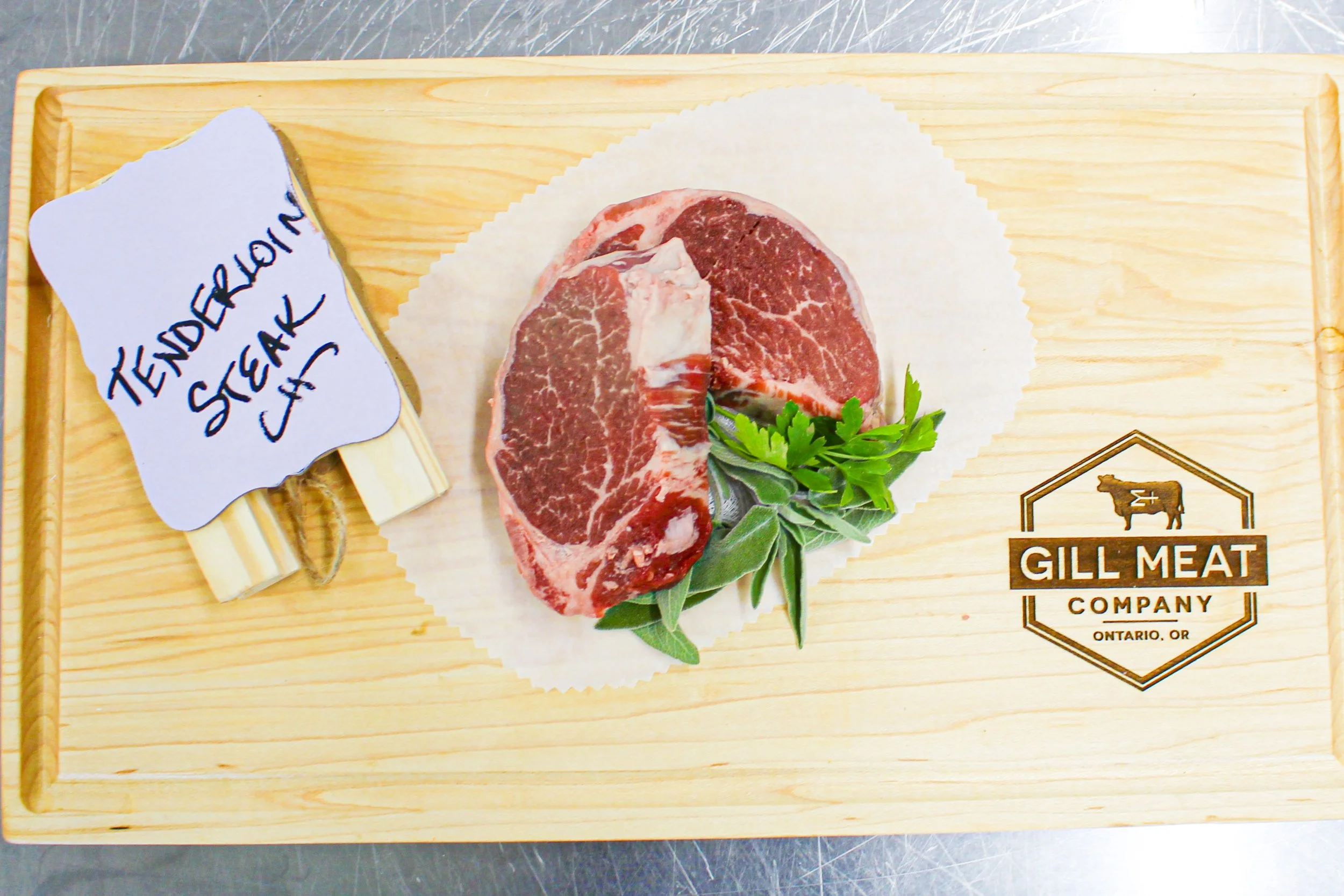 Raw T-bone steak from local beef on a wooden cutting board with a sprig of green herbs, labeled as 'Tenderloin Steak' with a lace ribbon on a small bow, and the 'Gill Meat Company' logo with a cow silhouette and 'Ontario, OR' written underneath.