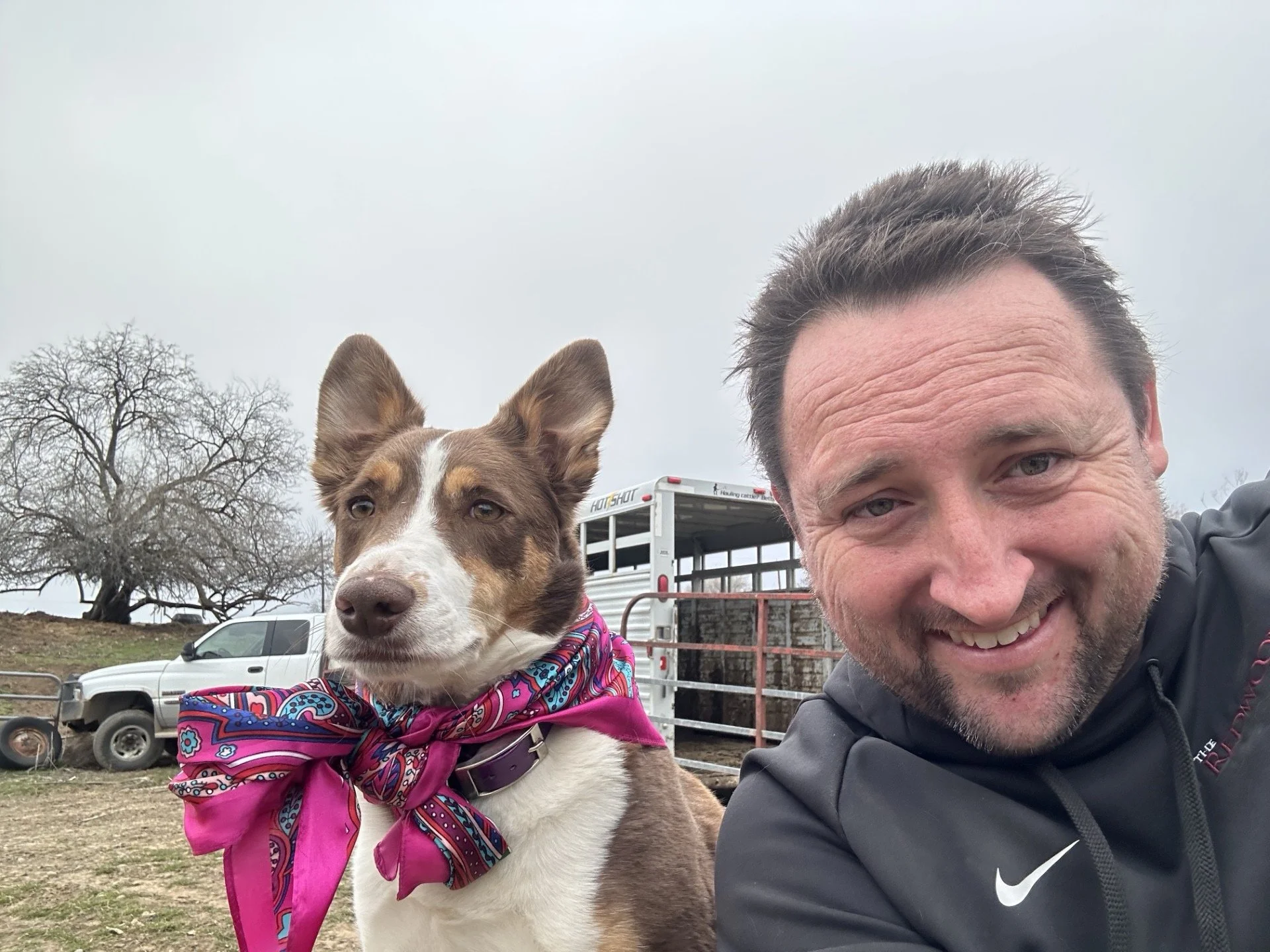 Ontario rancher smiling with a brown and white ranch dog wearing a pink bandana, outdoors with vehicles and a leafless tree in the background.