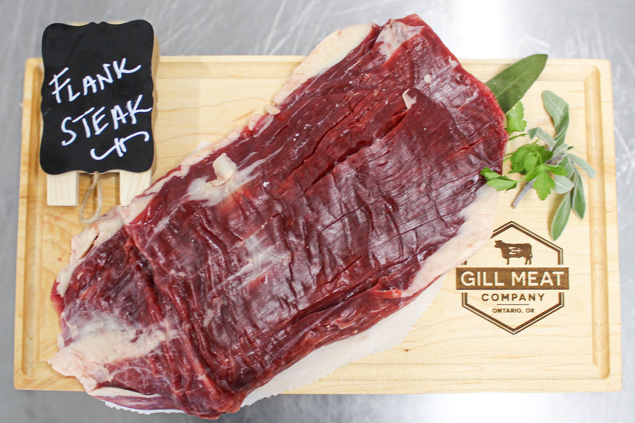 A large cut of raw beef flank steak on a wooden cutting board, labeled with a small chalkboard sign reading 'Flank Steak' and garnished with green herbs. The cut is from Gill Meat Company, Ontario, OR.