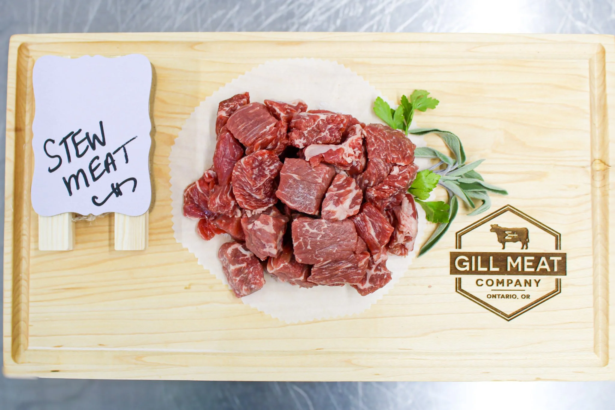 Raw stew meat on a wooden board with a small sign labeled 'Stew Meat', and some green herbs beside the meat, along with the 'Gill Meat Company' logo.