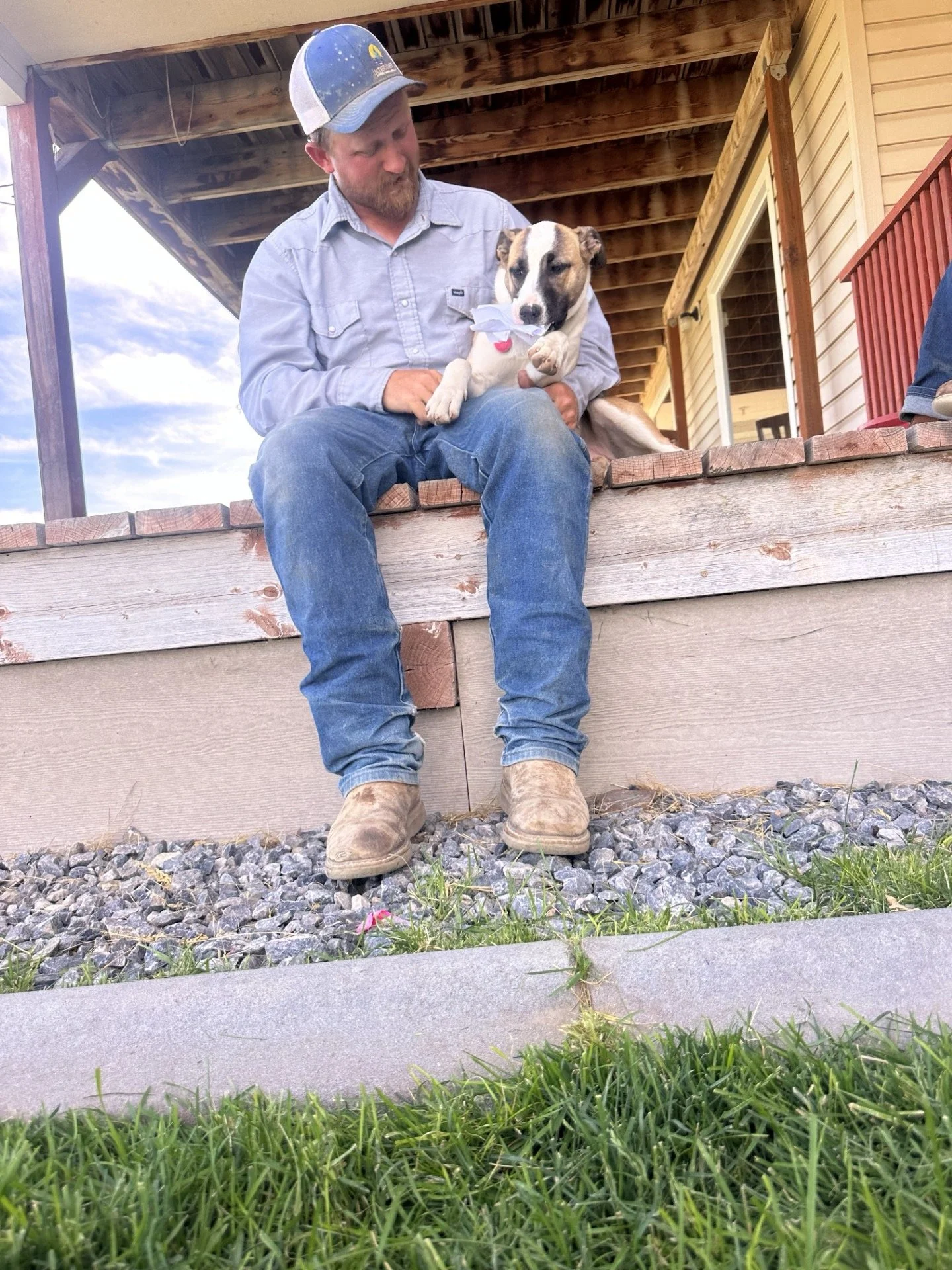 Farmer wearing a baseball cap, jeans, and boots sitting on a Treasure Valley farm front porch ledge holding a dog with a paper in its mouth, outside of a house with a wooden porch and railing.