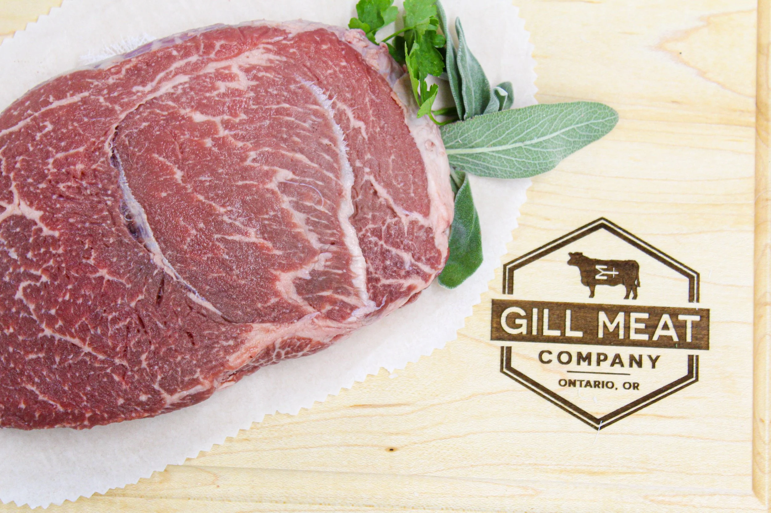 Raw grass fed corn finished well marbeled beef steak with green herbs on a wooden surface and Gill Meat Company logo.