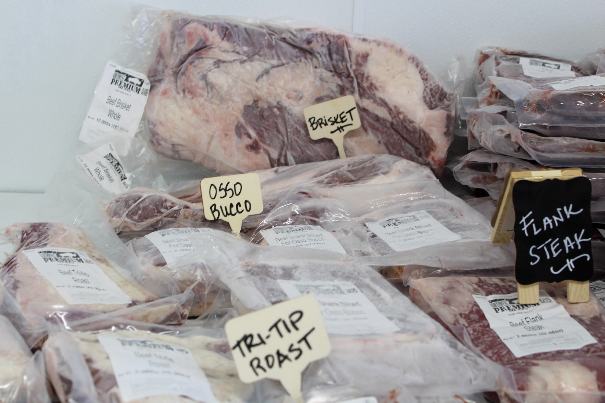 Various cuts of raw beef, including flank steak, tri-tip roast, and brisket, packaged in plastic and labeled with handwritten tags.