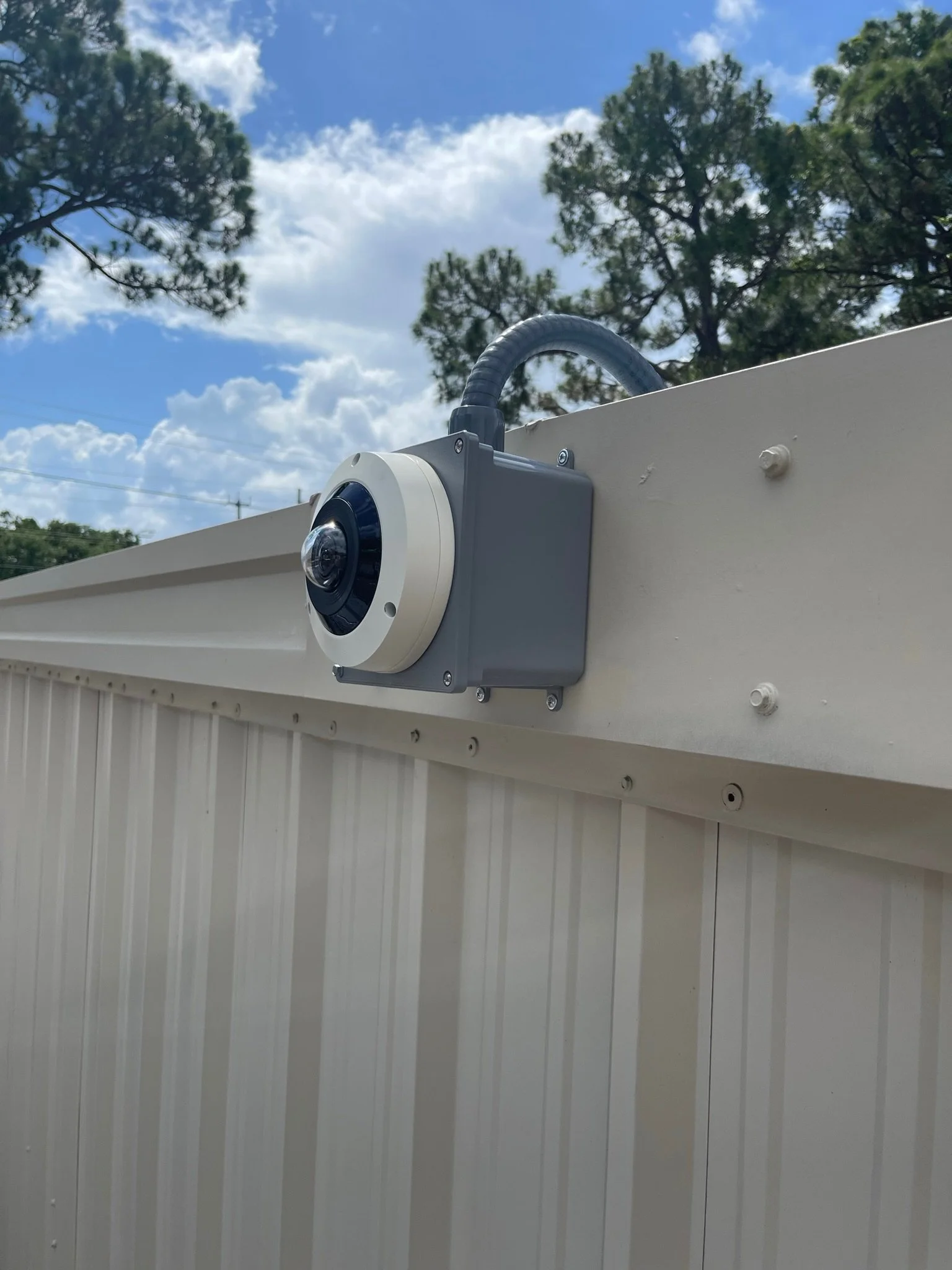 Close-up of a security camera mounted on a beige metal building with trees and a partly cloudy sky in the background.