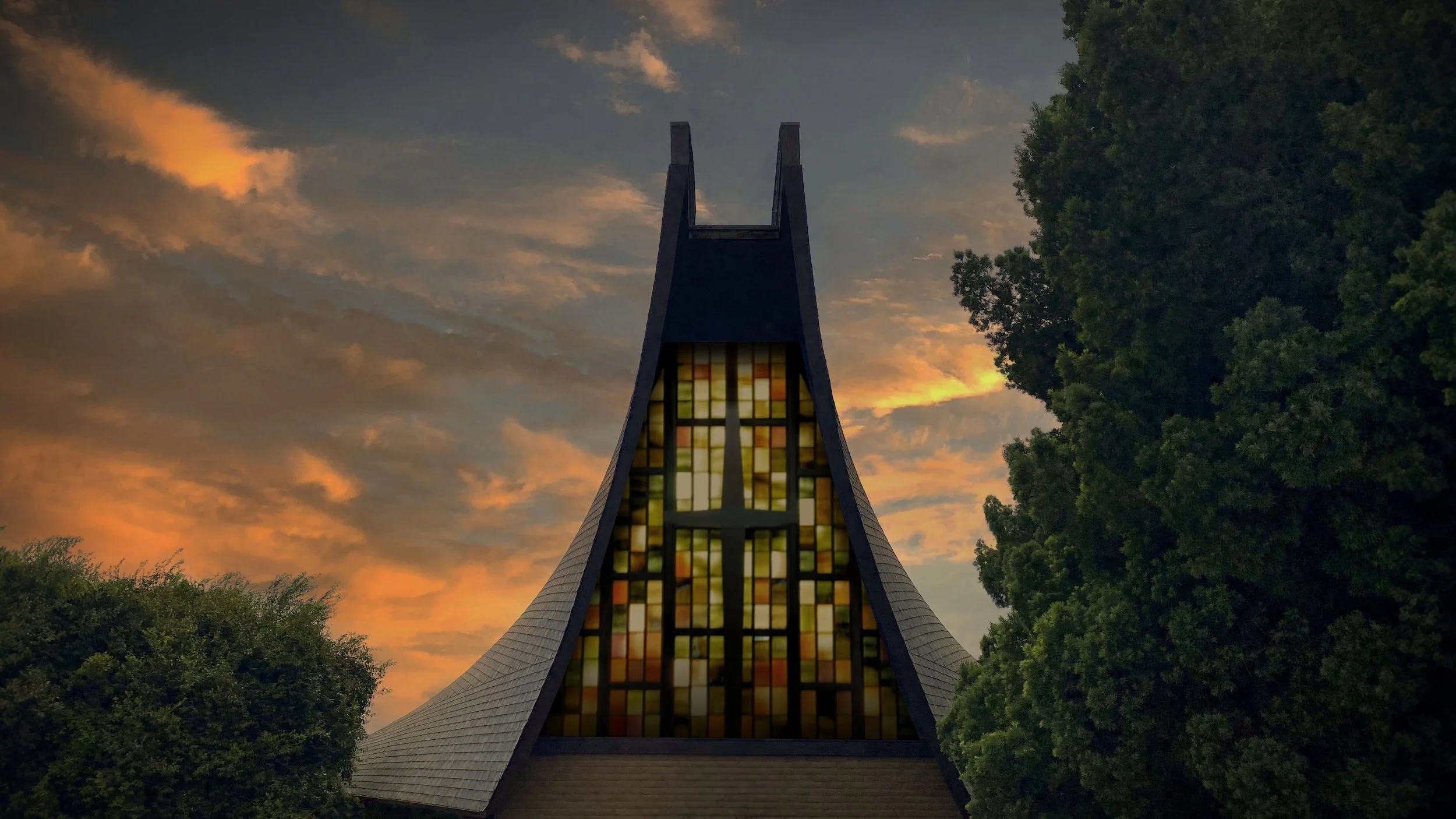 A church with modern architecture featuring a tall, pointed steeple and stained glass windows, set against a sunset sky with clouds and surrounded by green trees.