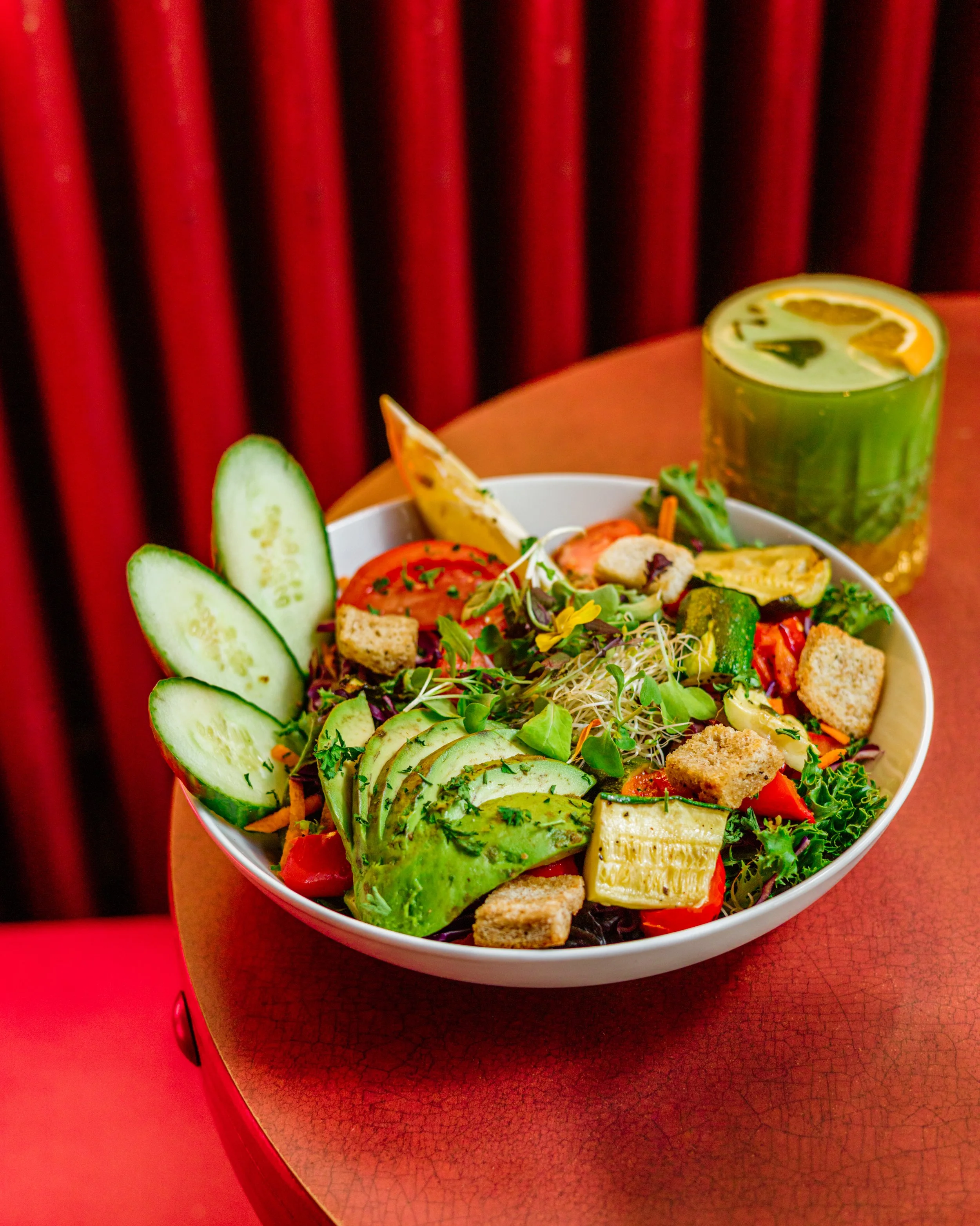 Une assiette de salade composée avec des tranches de concombre, avocat, tomates, croutons de pain, des légumes et des micro-pousses, accompagnée d'un verre de boisson citronnée au fond.