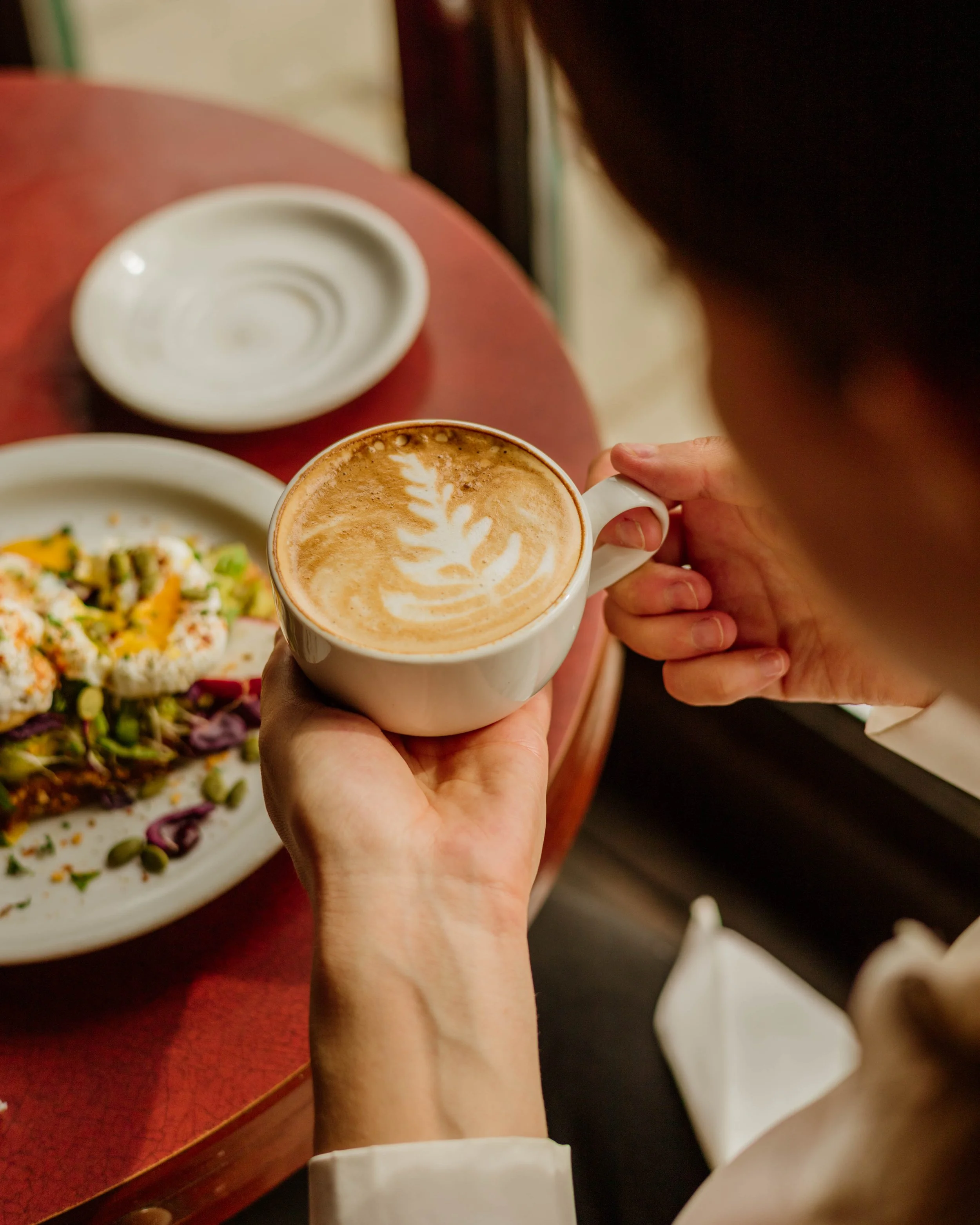 Une personne tenant une tasse de café avec un motif latte art, devant une assiette de nourriture dans un restaurant.