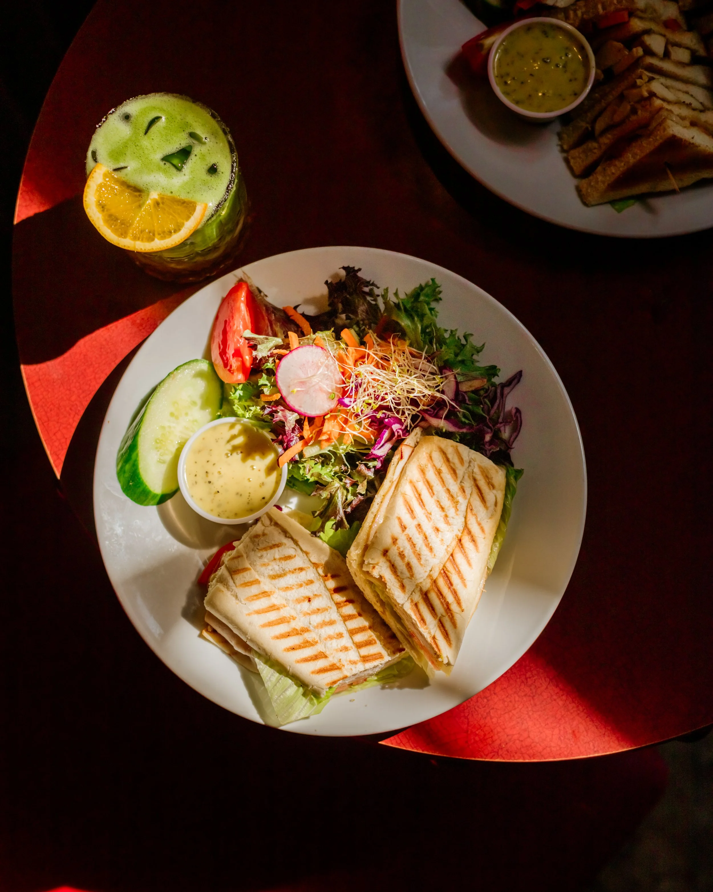 Assiette avec sandwich grillé, salade composée, concombre, carotte, radis, et sauce, accompagnée d'une boisson verte citronnée avec glaçons.