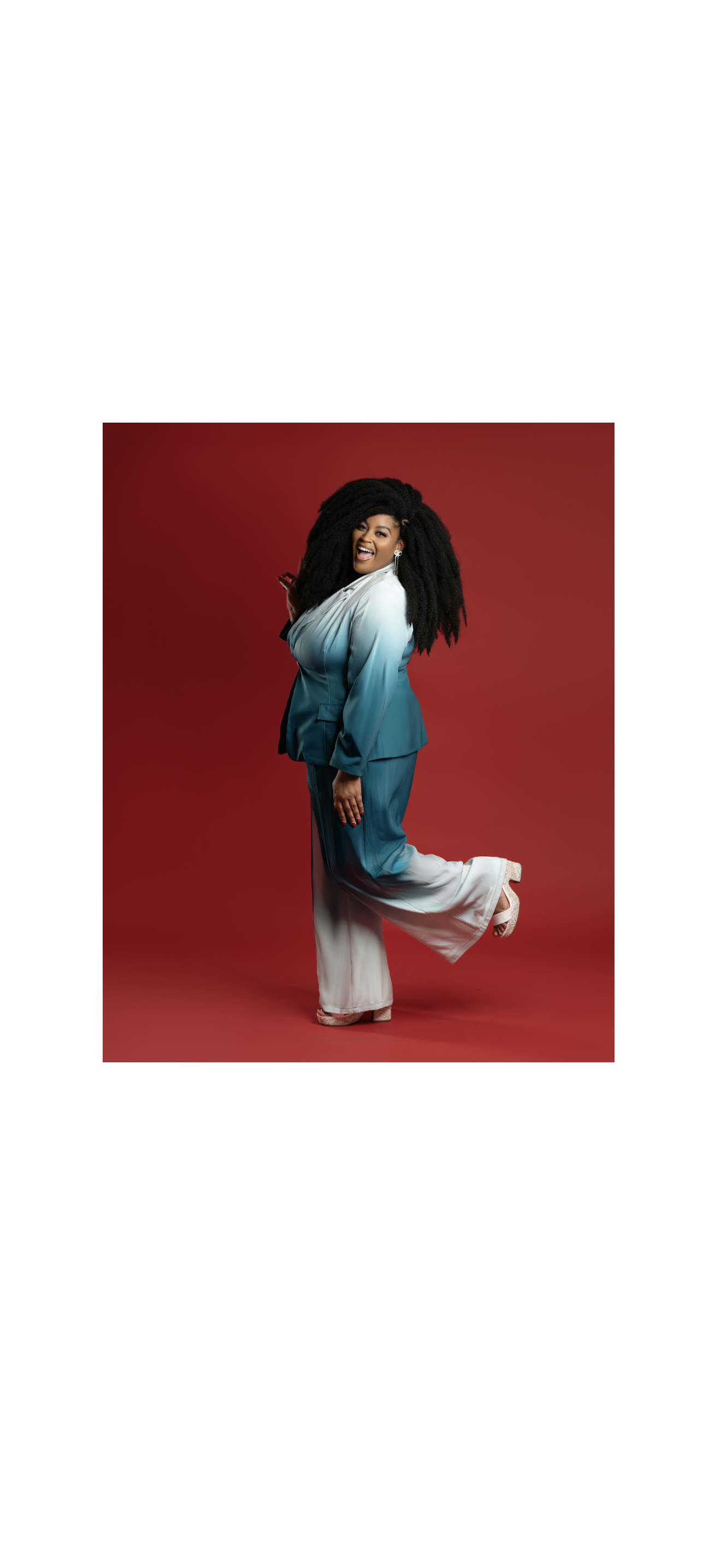 A woman with large black curly hair laughing, wearing a gradient blue and white satin suit with wide-legged pants, standing on one leg against a red background.