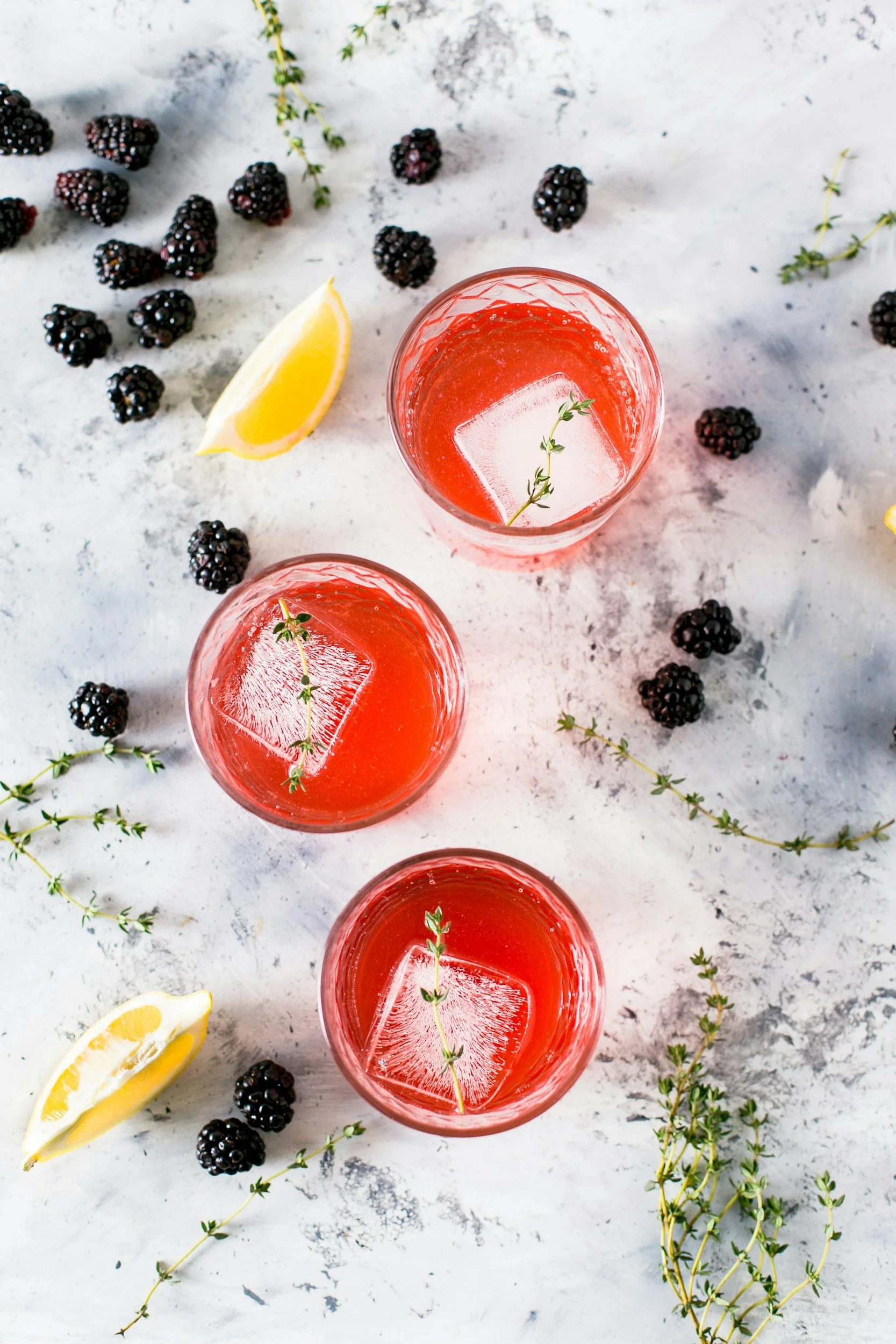 Three glasses of pink beverage with ice and thyme, garnished with lemon wedges, and surrounded by blackberries and sprigs of thyme on a white marbled surface.