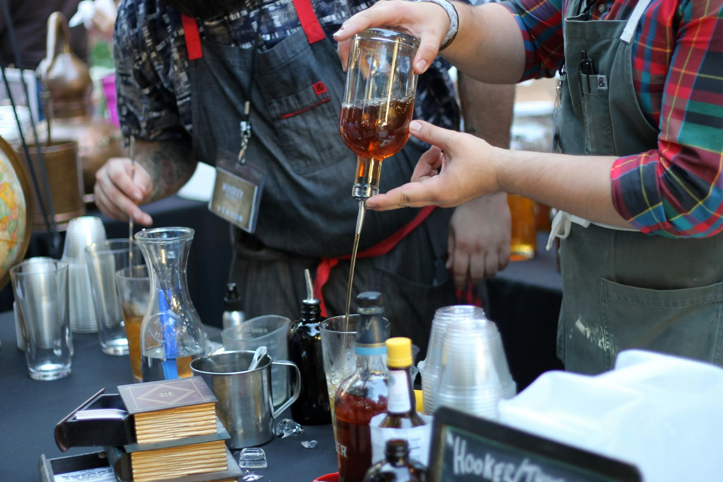 People at a table with glassware, bottles, and various tools, brewing or mixing a beverage, possibly at a craft event or workshop.