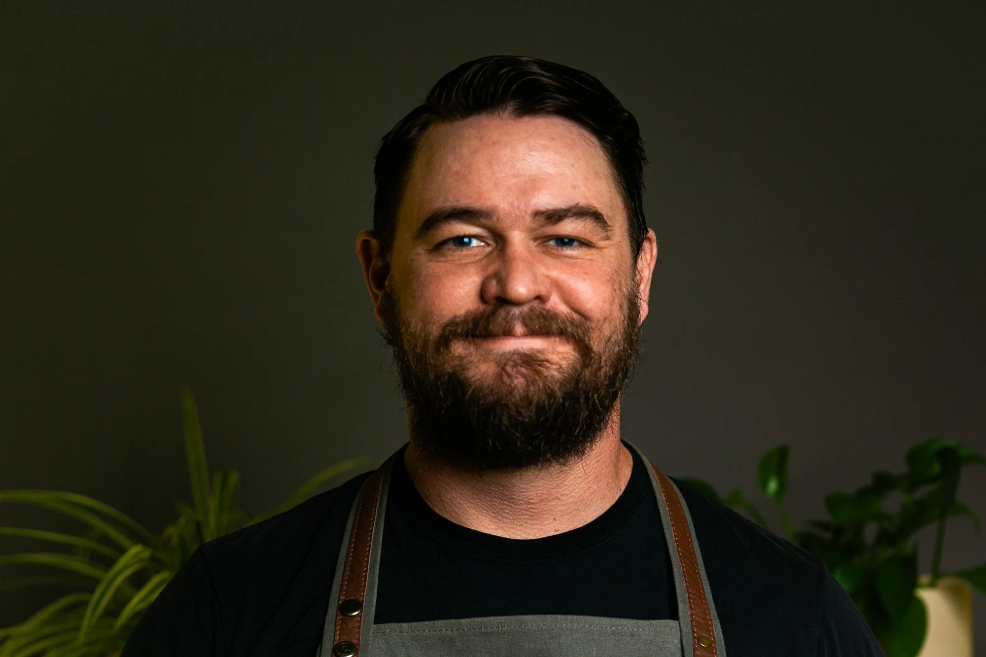 Portrait of a man with dark hair, a beard with some gray, and blue eyes, smiling, standing in front of green plants on a dark background.