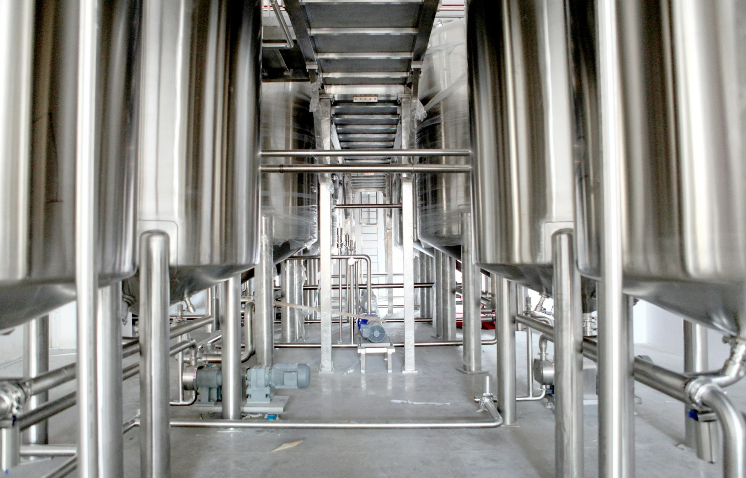View of an industrial facility with stainless steel tanks and pipelines, viewed from ground level looking down a corridor between the tanks, with a staircase in the background.