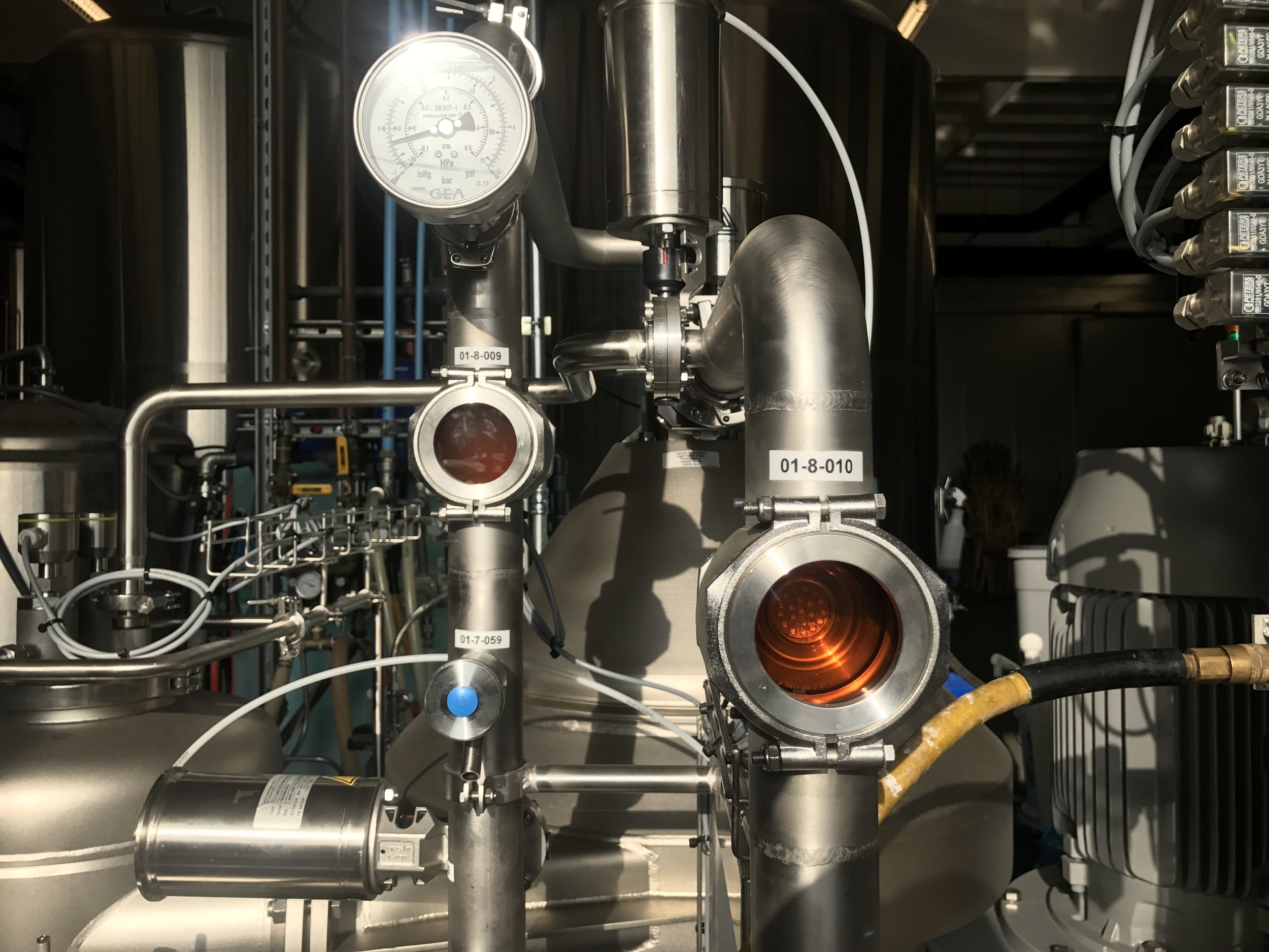 Close-up of industrial machinery with pipes, gauges, and valves, including a pressure gauge at the top and illuminated orange light inside a pipe.