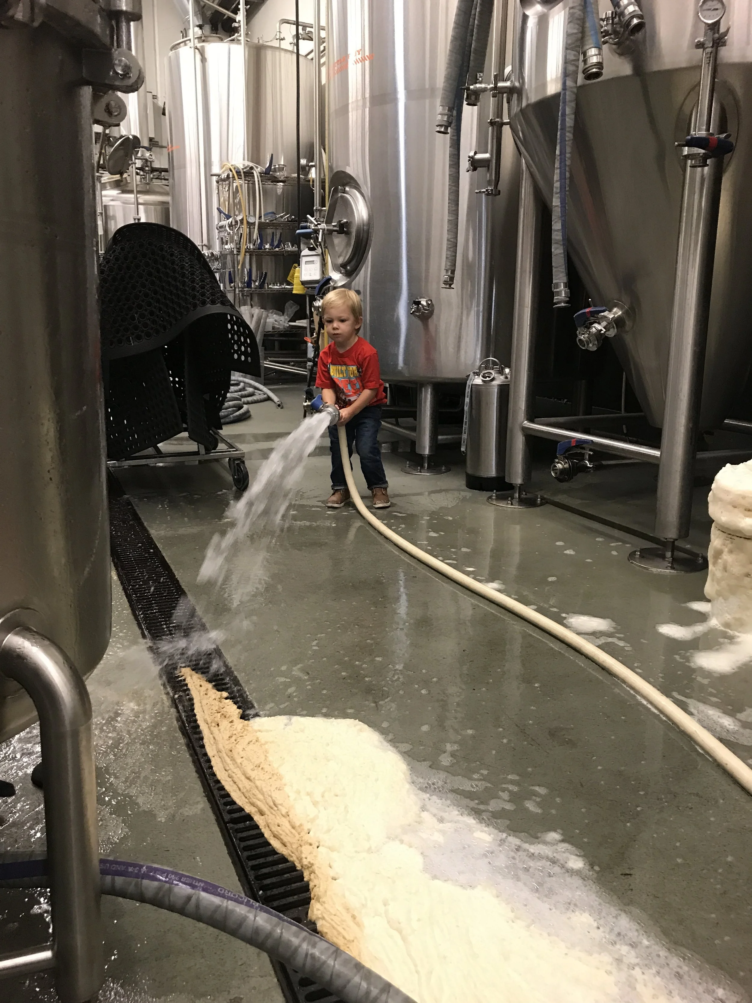 A young boy in a red shirt is using a hose to spray water in a brewery or industrial beverage production room, surrounded by large stainless steel tanks and brewing equipment.